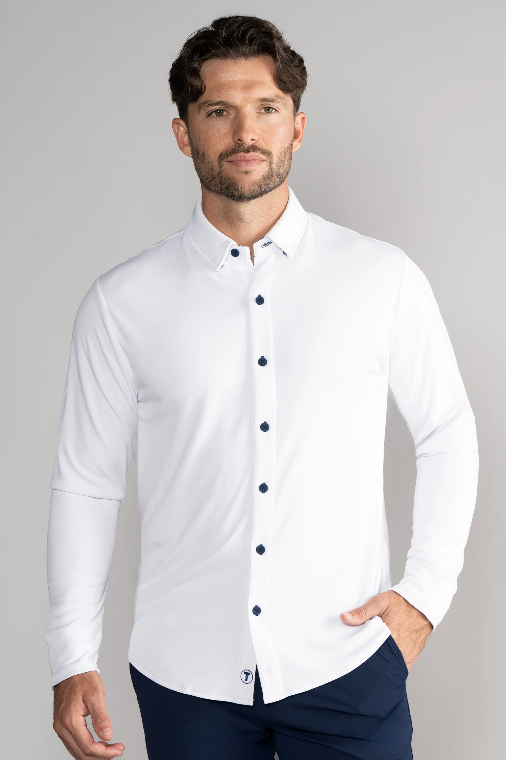 A man with short, wavy brown hair and a trimmed beard wears the Twillory Long Sleeve Button Down Polo Contrast in White Navy, dark blue pants, and stands against a light gray background with one hand in his pocket and a neutral expression.