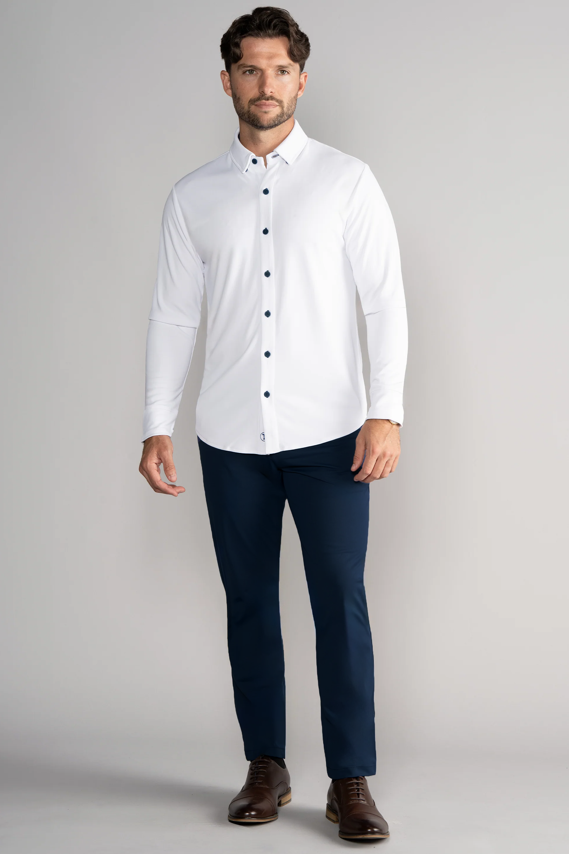 A man with short brown hair and a trimmed beard stands against a light gray background, wearing the Twillory Long Sleeve Button Down Polo Contrast in White Navy. He looks slightly to the right, styled with navy pants and brown dress shoes.