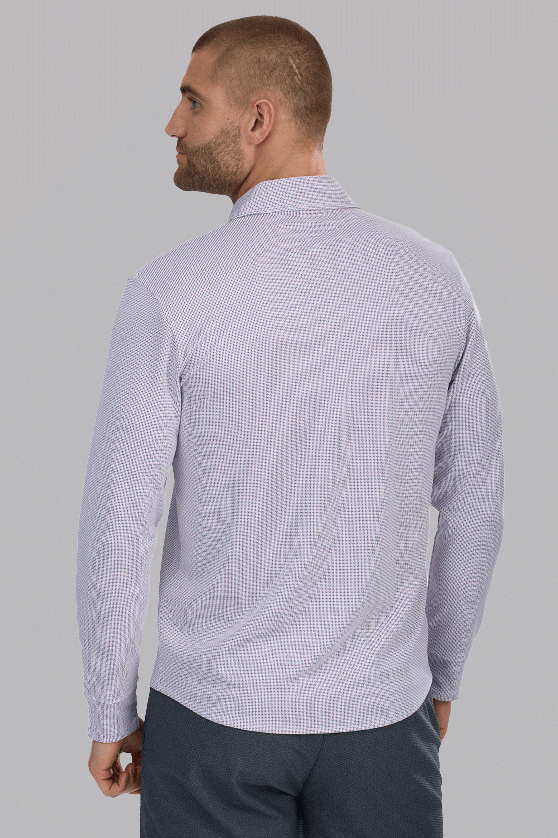 A man with short hair and a beard is seen from the back, wearing the Twillory Long Sleeve Button Down Polo Dot in Orange Blue. He stands against a plain light gray background, looking over his left shoulder.