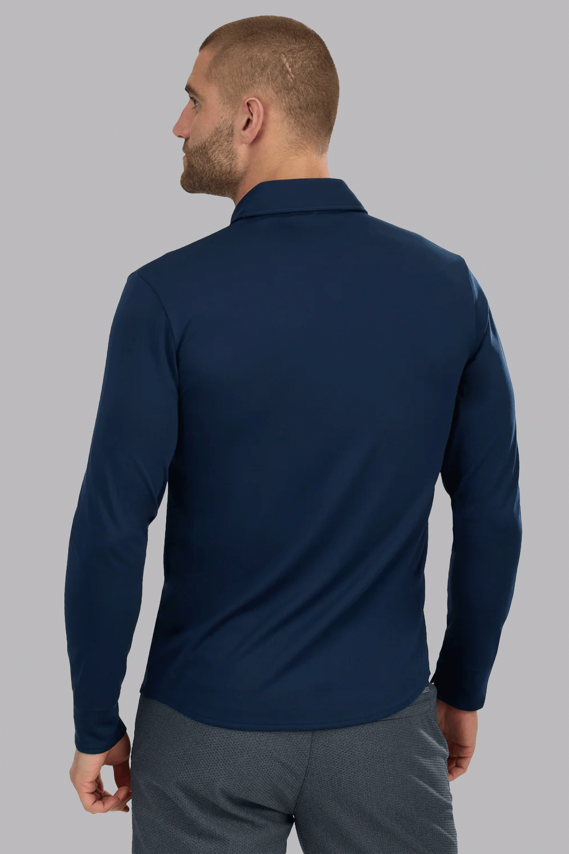 A man is seen from the back, wearing the Twillory Long Sleeve Button Down Polo Contrast in Navy White. He stands against a plain, light gray background, looking slightly left with his arms relaxed by his sides.
