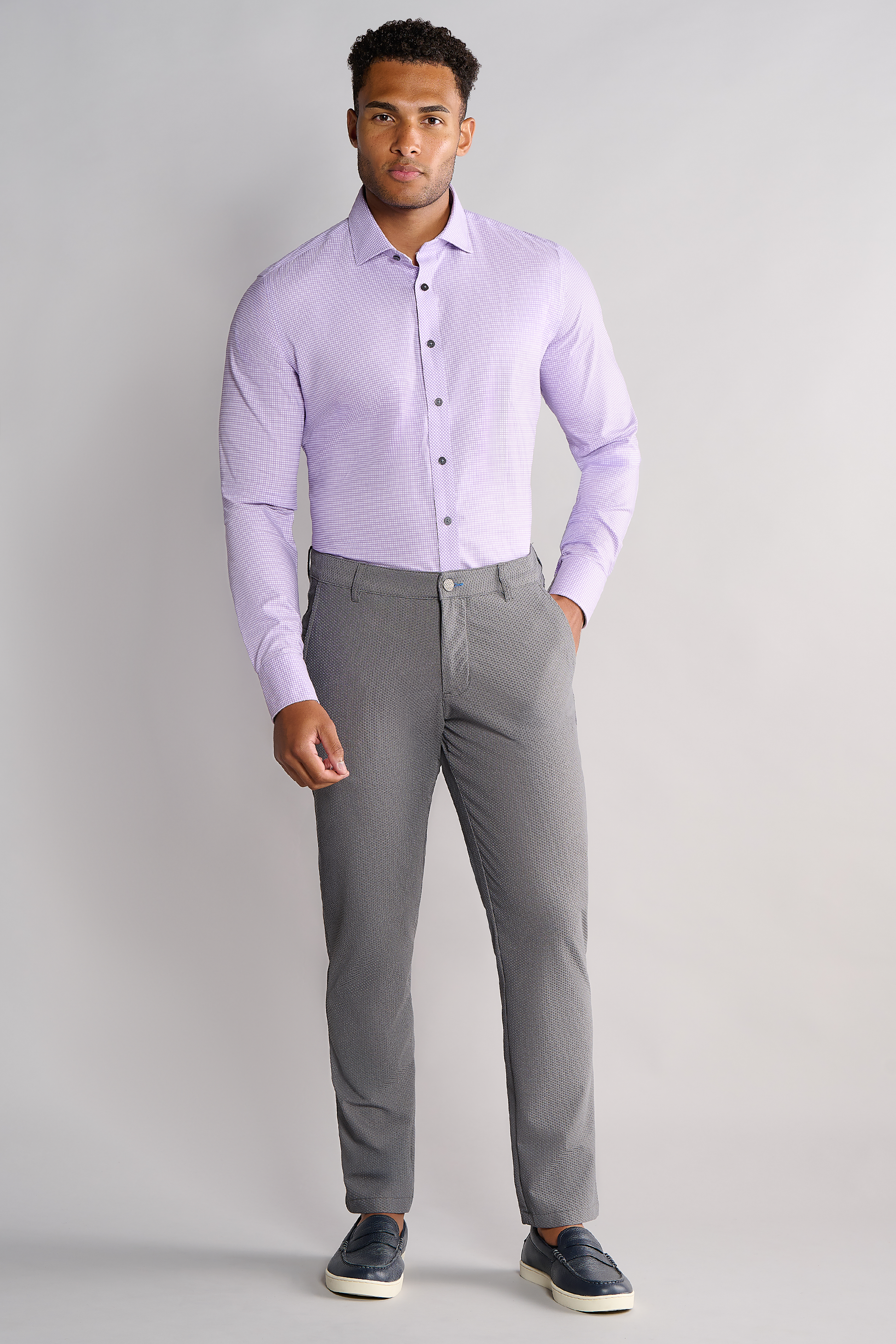A man stands against a light gray background, wearing Twillory's Leader Check // Lavender performance shirt, tucked into slim-fit gray trousers. He has one hand in his pocket and wears dark slip-on shoes with white soles, looking forward neutrally.