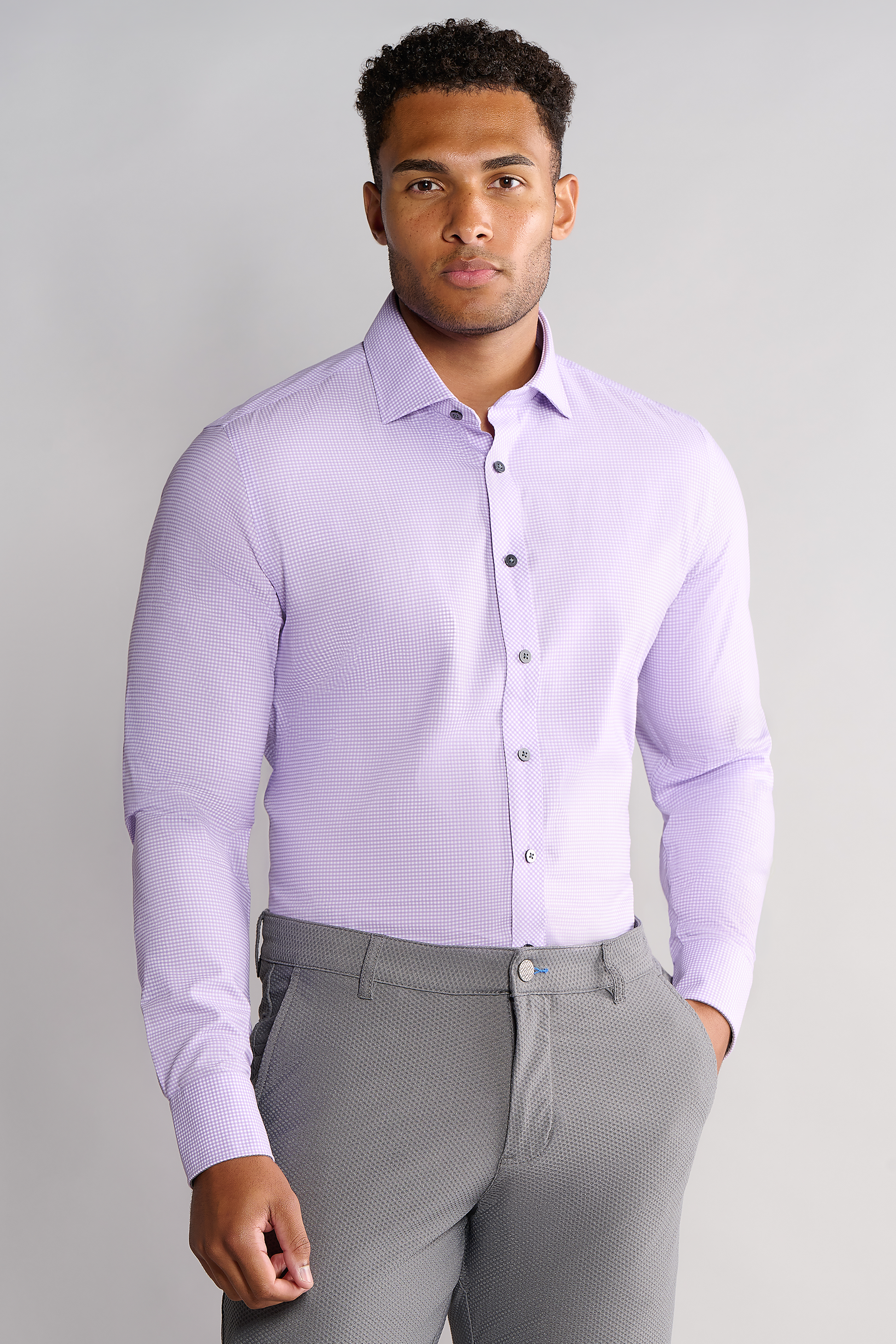 A man faces forward, wearing Twillory’s Leader Check // Lavender shirt with 4-way stretch and gray dress pants. He has short curly hair, a trimmed beard, relaxed hands at his sides, and stands against a plain light gray background.