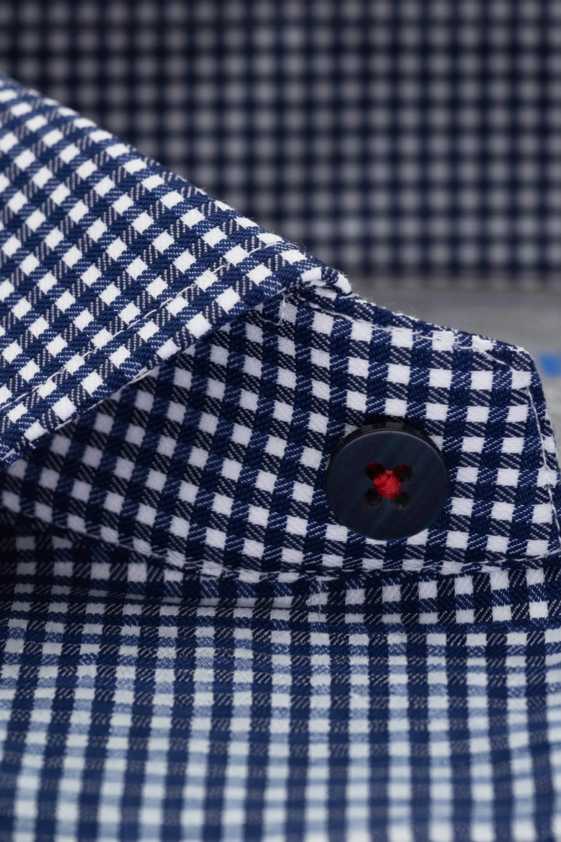 A close-up of Twillory’s Leader Check // Blue shirt collar, featuring blue and white checks, a black button with red stitching, and a textured checkered background that highlights its moisture-wicking fabric.