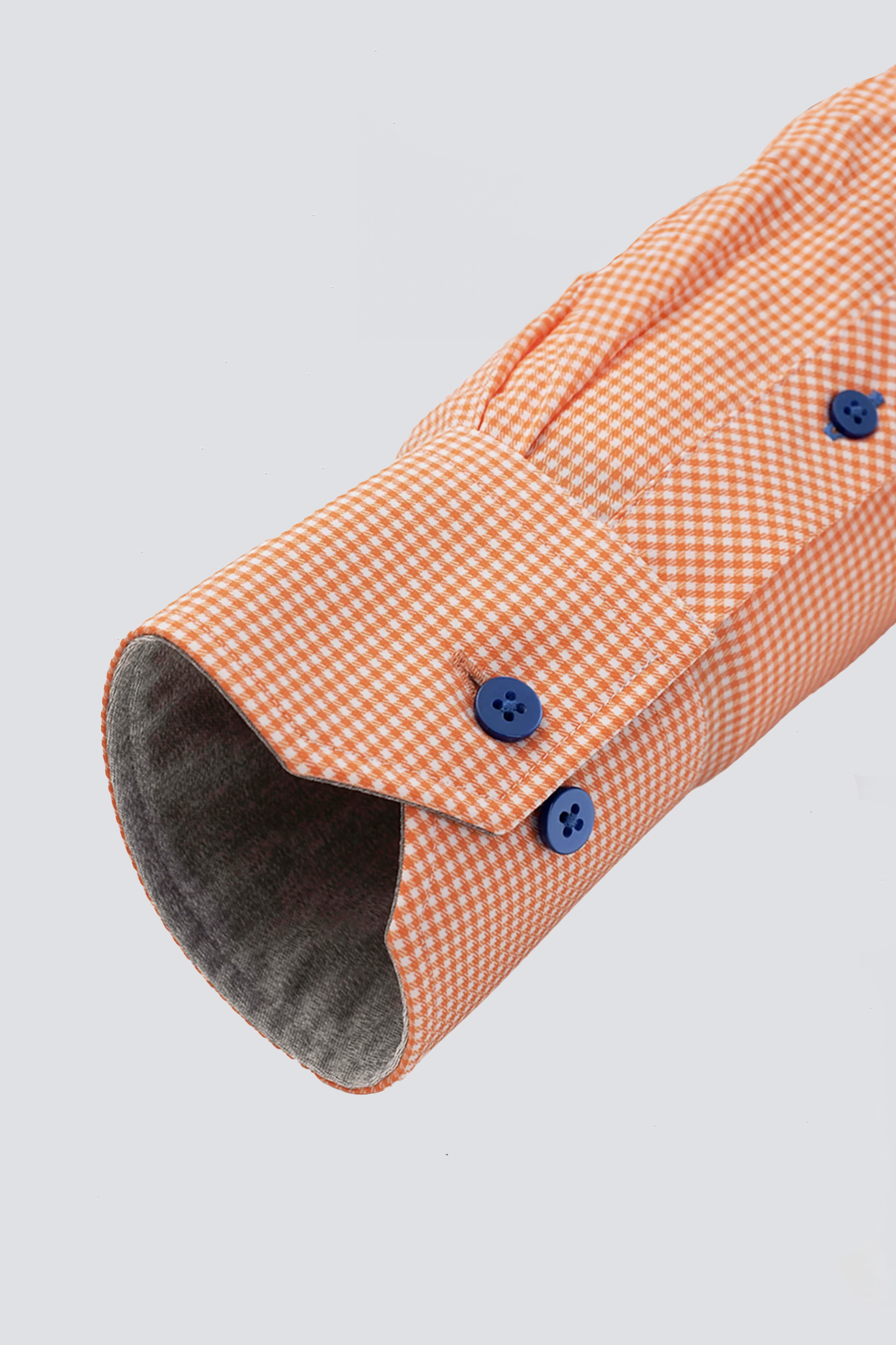 A close-up of the Twillory Leader Check // Orange Performance Shirt sleeve shows a pointed cuff with two blue buttons, a grey inner lining, and an orange and white small check pattern with 4-way stretch. The background is plain light grey.