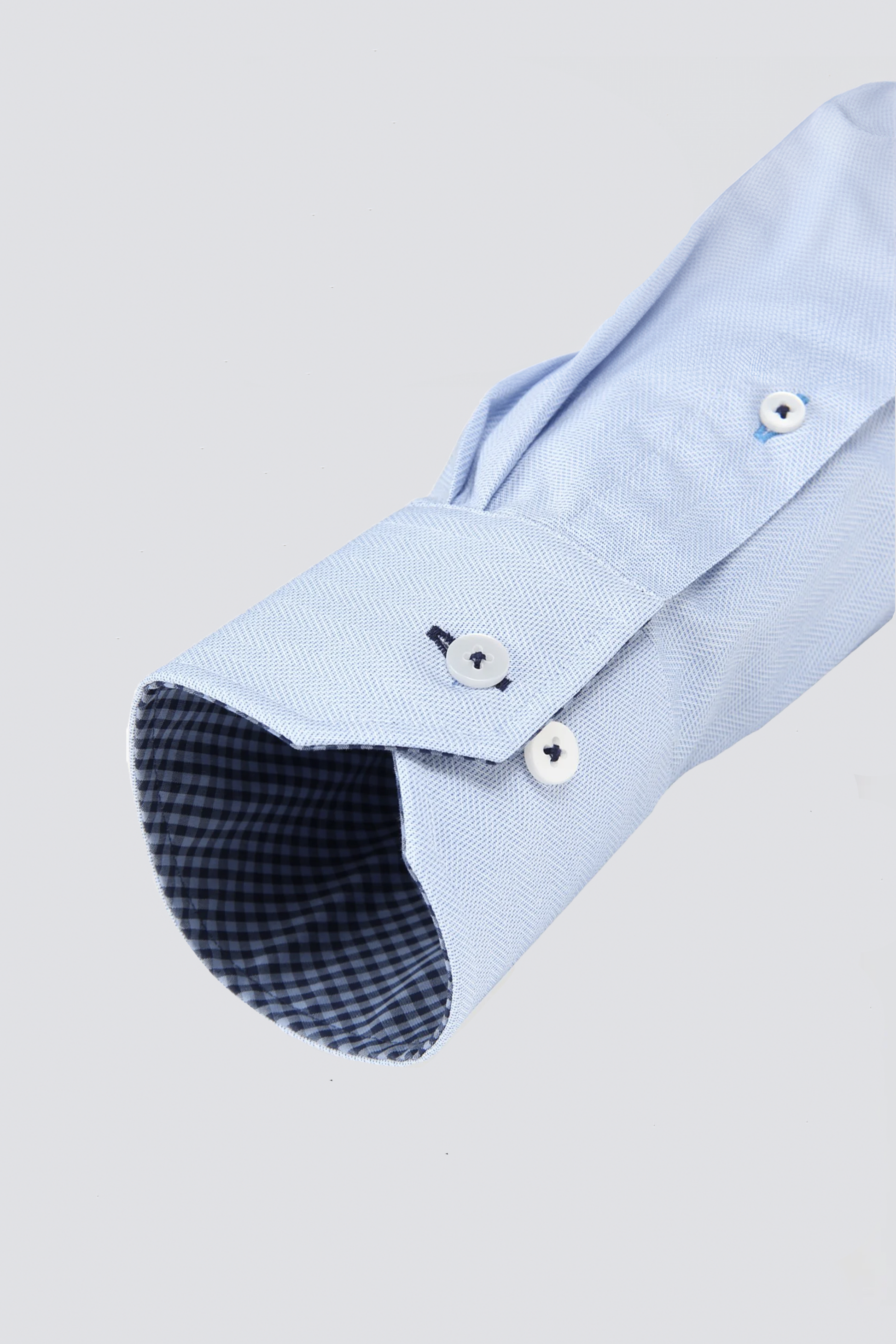 A close-up of the Twillory Leader Contrast // Blue Check performance shirt's light blue herringbone sleeve, buttoned with two white buttons and featuring 4-way stretch plus contrasting dark blue and white check inside the cuff. Light gray background.