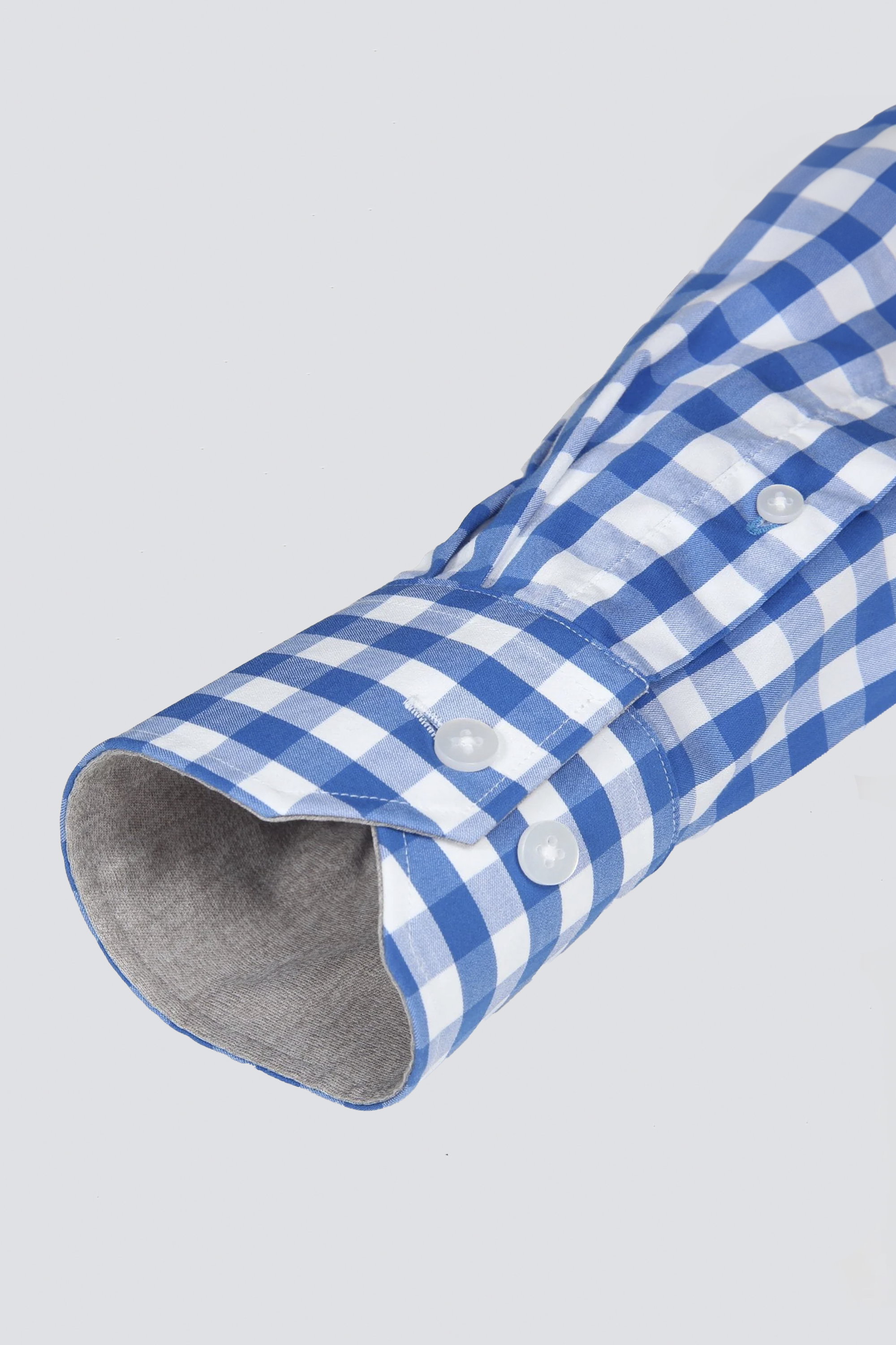 A close-up of Twillory's Leader Gingham // Blue White long-sleeve performance shirt shows its blue and white check pattern, white buttons, unbuttoned cuff with light gray lining, and moisture-wicking fabric against a plain background.