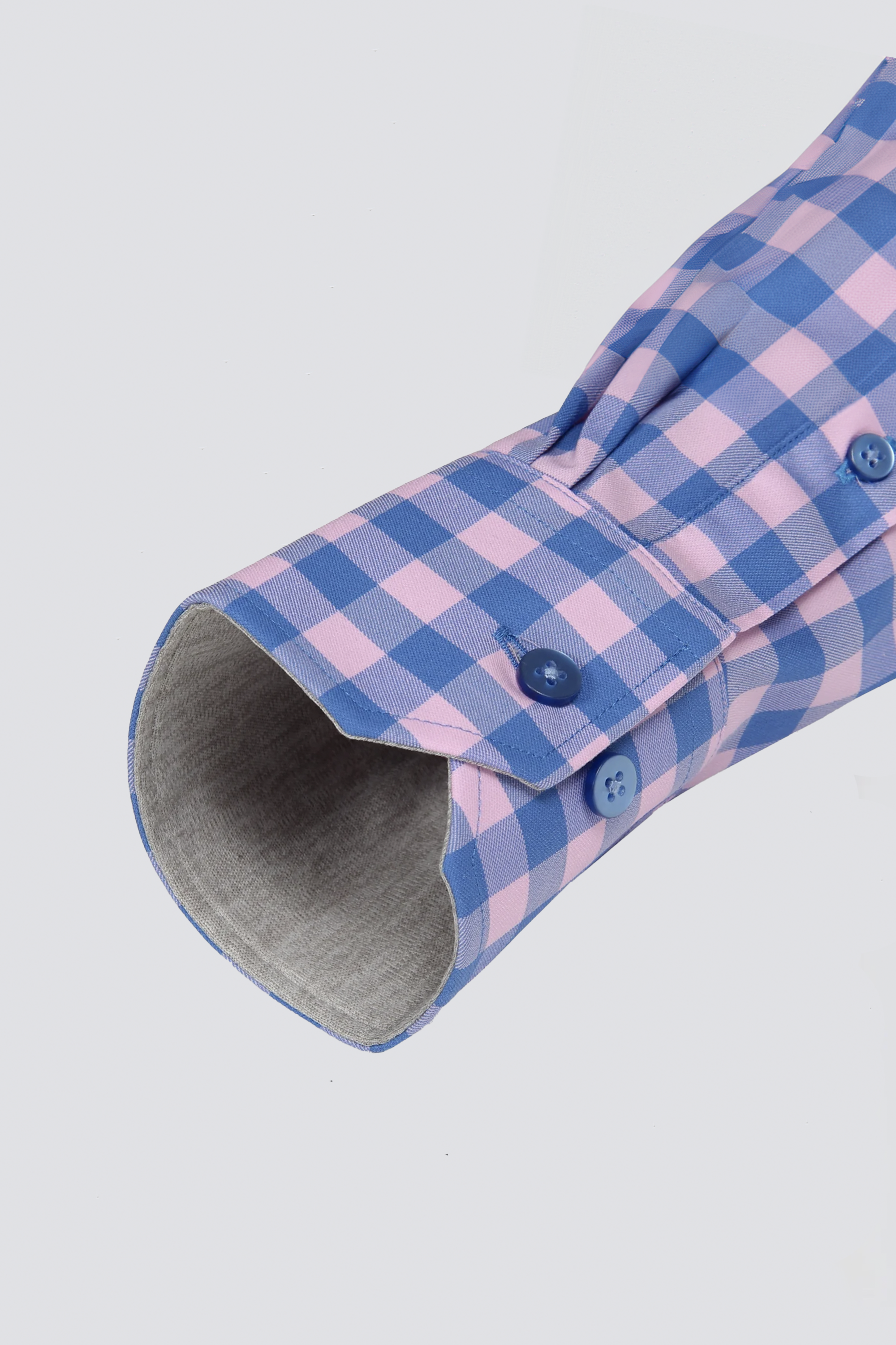 A close-up of the Twillory Leader Gingham // Pink Blue shirt cuff shows blue and pink checkered fabric, two blue buttons, a visible buttonhole, soft gray inner lining, and a neat press against a plain background.