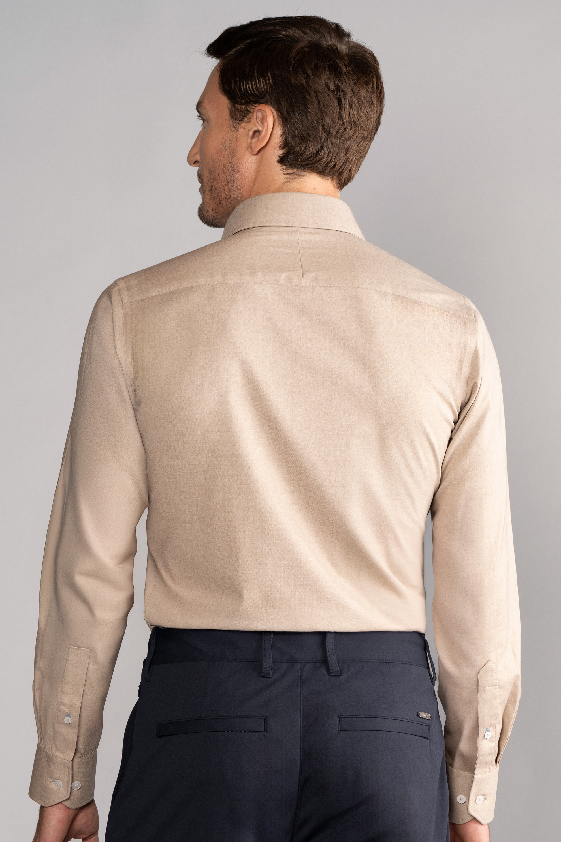 A man stands with his back to the camera, wearing Twillory’s Leader Solid // Heather Sepia performance shirt tucked into dark navy dress pants. His styled short brown hair contrasts against a plain light gray background.