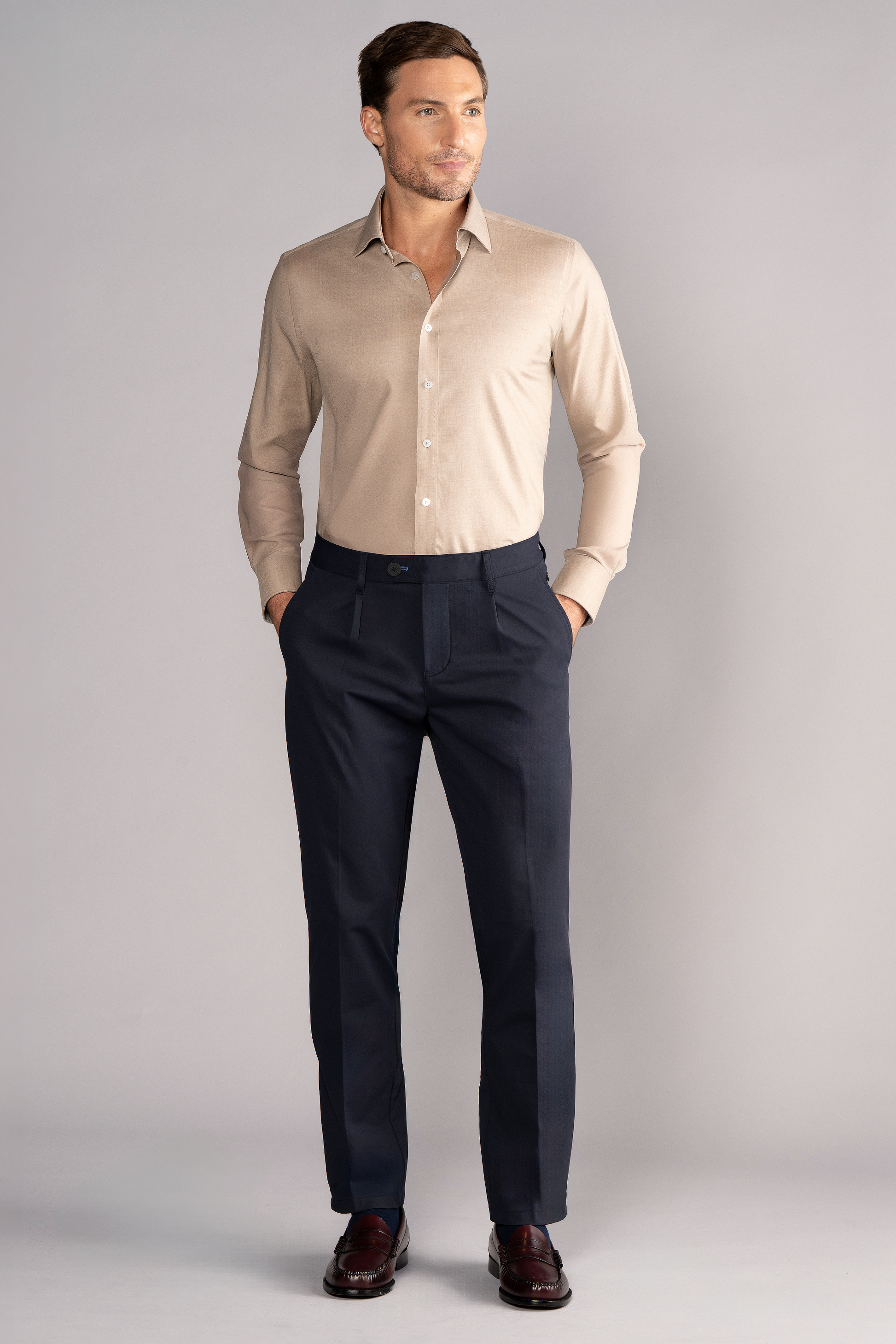 A man with short brown hair and a trimmed beard stands facing forward against a gray background, wearing Twillory’s Leader Solid // Heather Sepia performance shirt tucked into dark navy dress pants with a belt, and dark brown dress shoes. Hands in pockets.