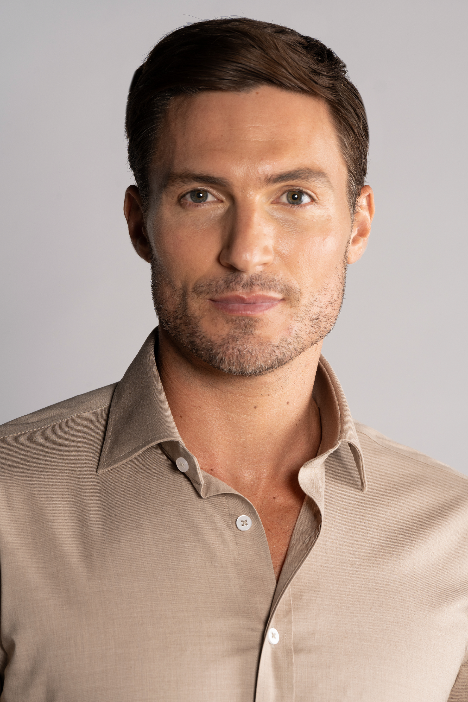 A man with short brown hair and light stubble, wearing the Twillory Leader Solid // Heather Sepia performance shirt with top button open, looks at the camera against a light gray background, showing a neutral, slight smile.