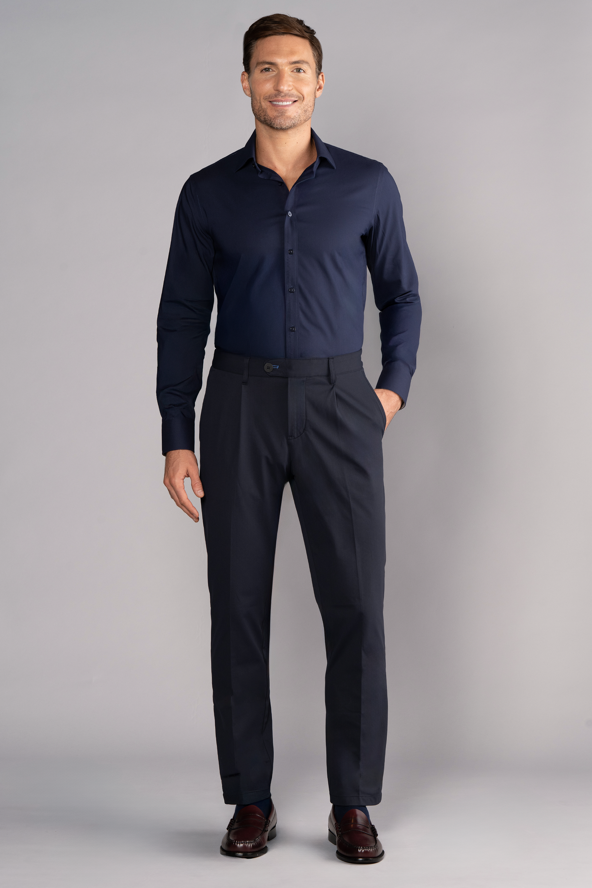A man smiles against a light gray background, wearing Twillory’s Leader Solid // Navy Performance Shirt and matching navy dress pants, tucked in neatly. His right hand is in his pocket and he completes the formal look with dark brown dress shoes.