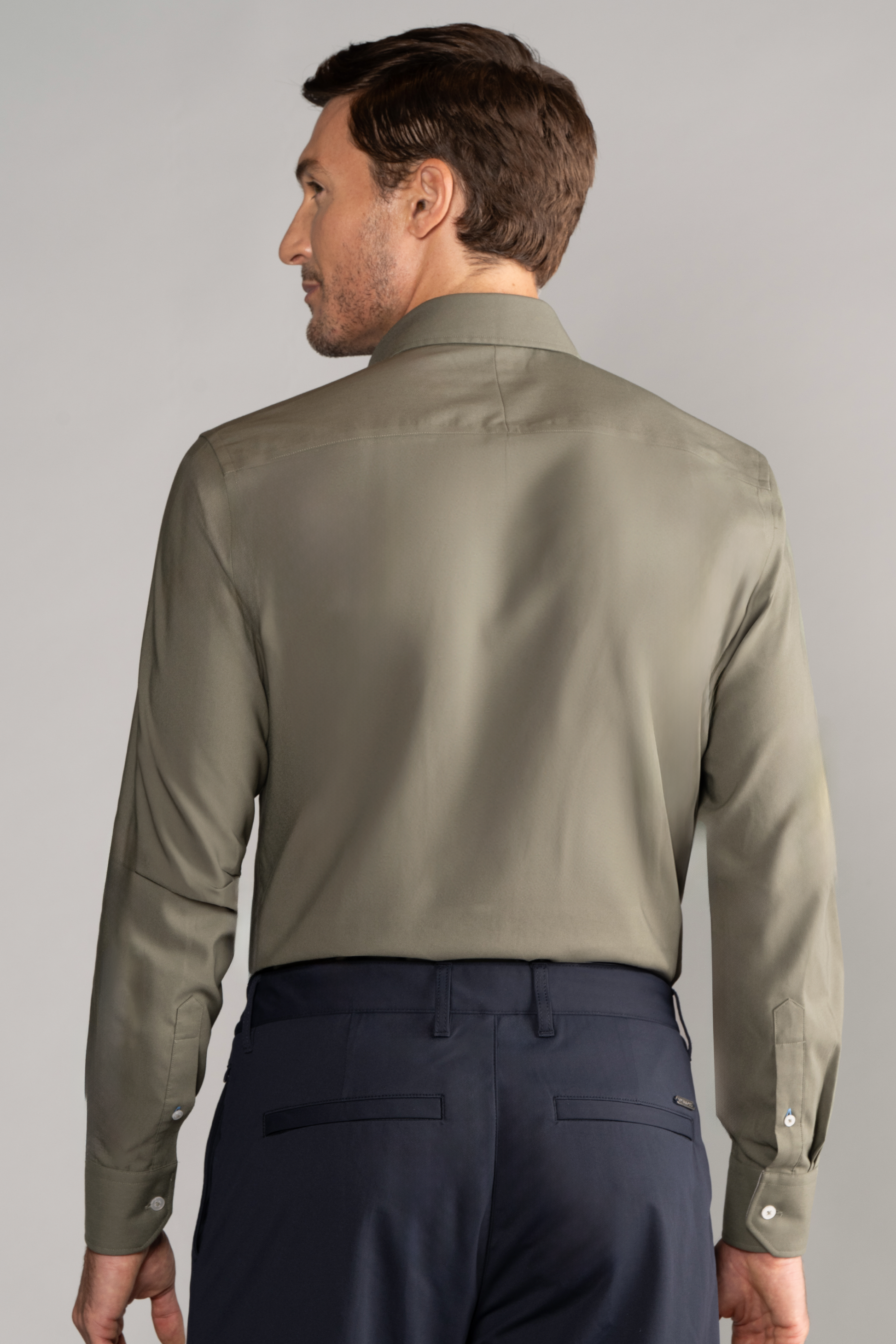 A man with short brown hair, seen from behind, wears the Twillory Leader Solid // Olive long-sleeve shirt and dark navy dress pants, standing against a plain light gray background.