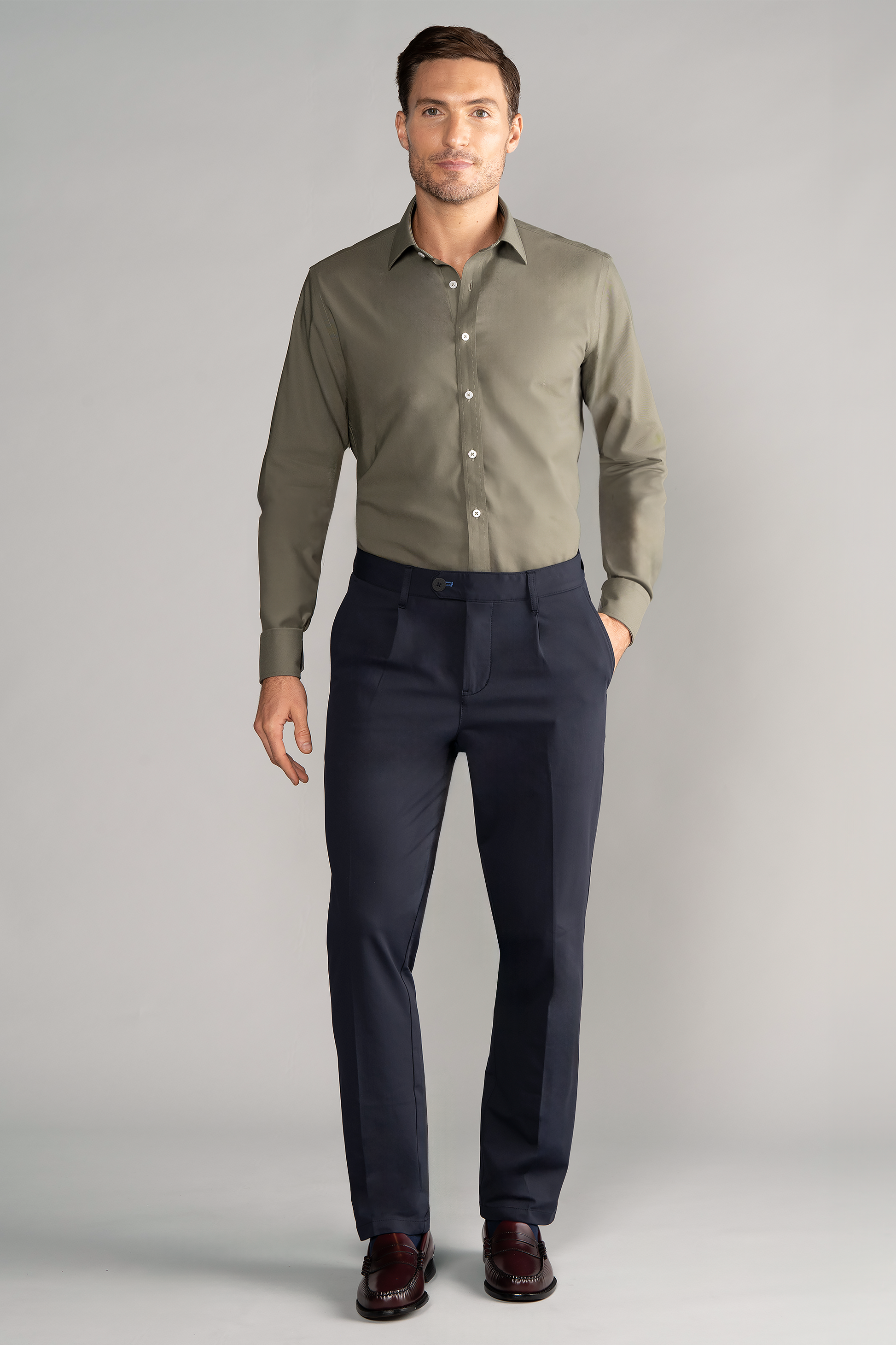 A man with short brown hair and a trimmed beard stands against a light gray background. He wears Twillory's Leader Solid // Olive performance shirt, dark navy dress pants, and brown dress shoes, with one hand in his pocket.