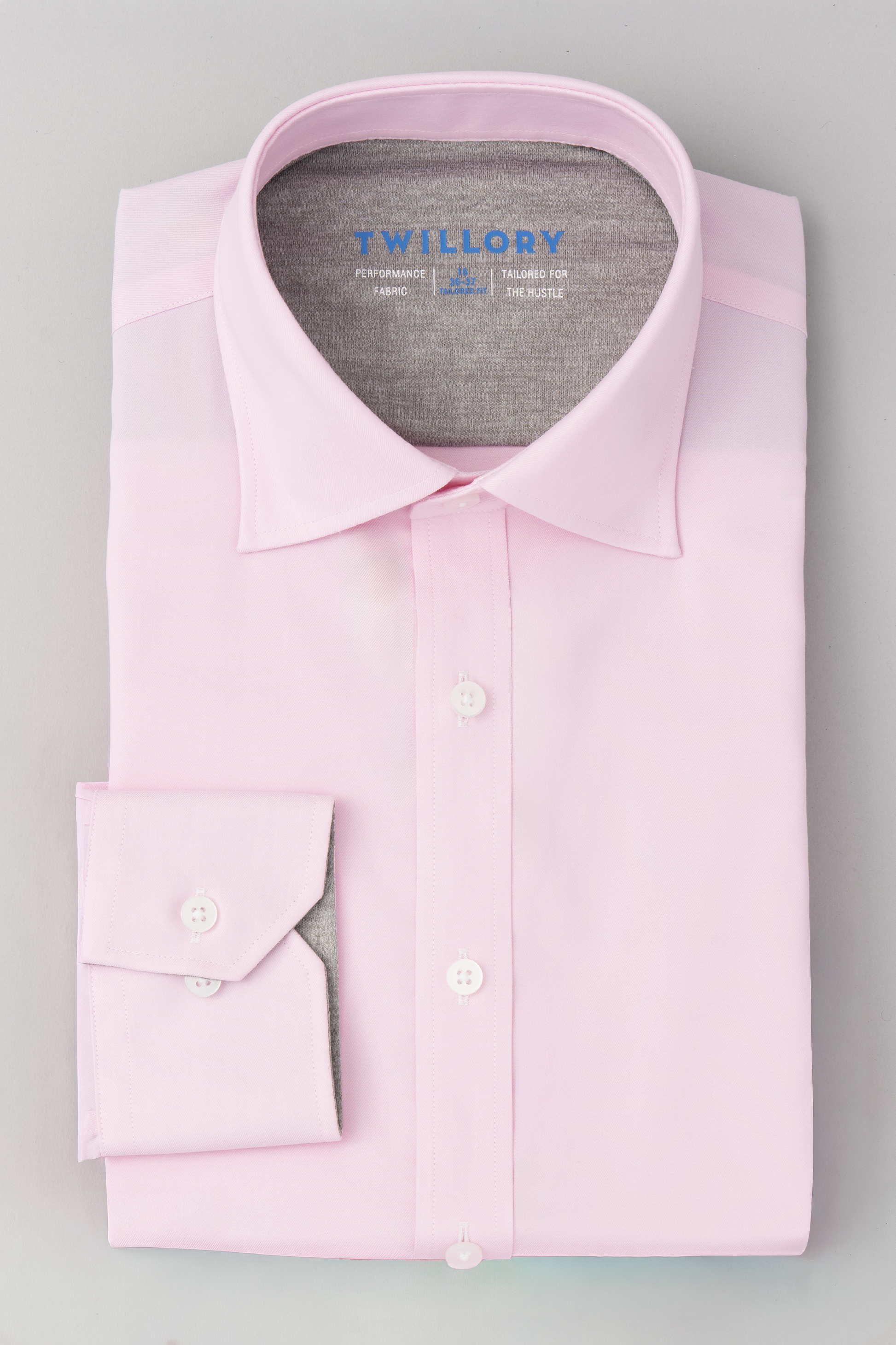 The Twillory Leader Solid // Pink shirt, featuring 4-way stretch, white buttons, and a classic collar, is neatly folded on a light gray surface with "Twillory" in blue on the inside collar. One cuff is visible.