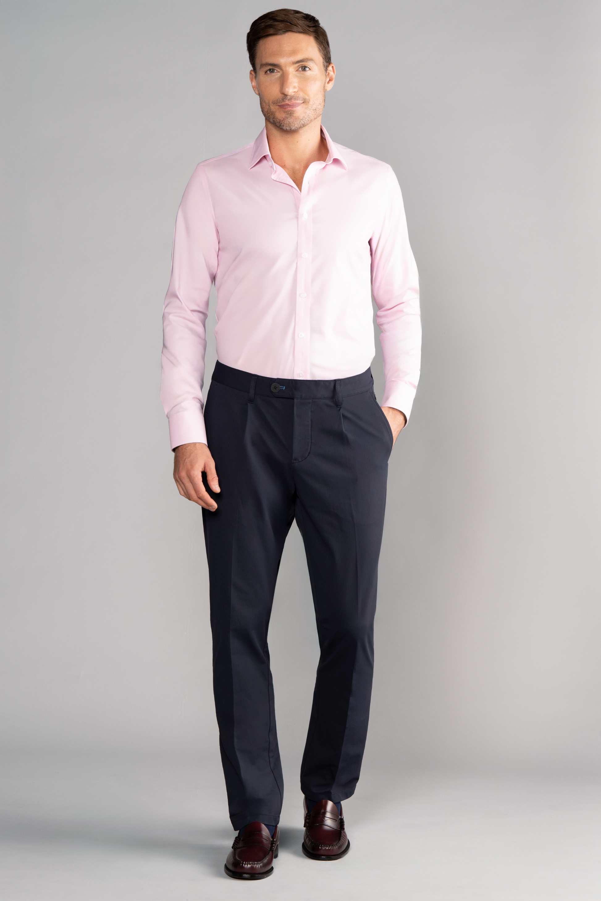 A man stands facing forward against a light gray background, wearing Twillory's Leader Solid // Pink performance shirt, dark navy dress pants, and dark brown dress shoes. His left hand is in his pocket; he has short brown hair and a neat beard.