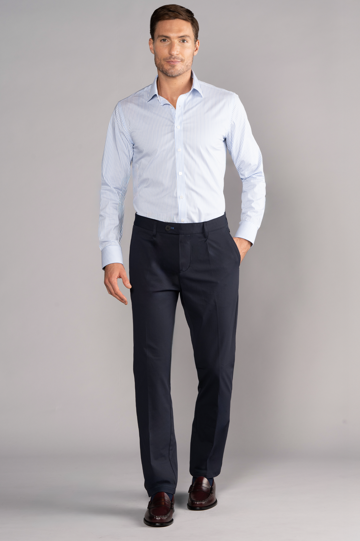 A man stands against a gray background, wearing Twillory’s Leader Stripe // Sky long-sleeve performance shirt tucked into dark navy dress pants, with dark brown dress shoes. He has short brown hair, a neutral expression, and one hand in his pocket.