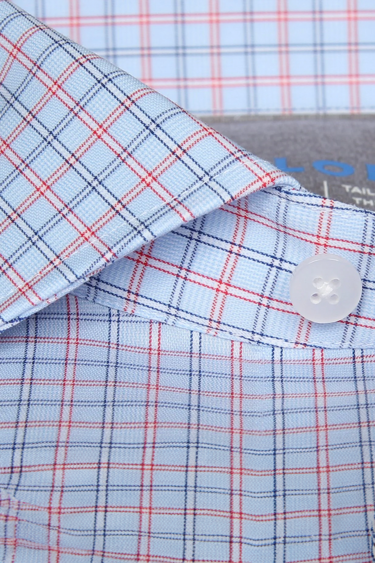 Close-up of the Twillory Leader Windowpane // Red Blue performance shirt, featuring a light blue base with red and navy plaid, crisp fabric, neat stitching, and a white button. A similar moisture-wicking shirt appears in the background.