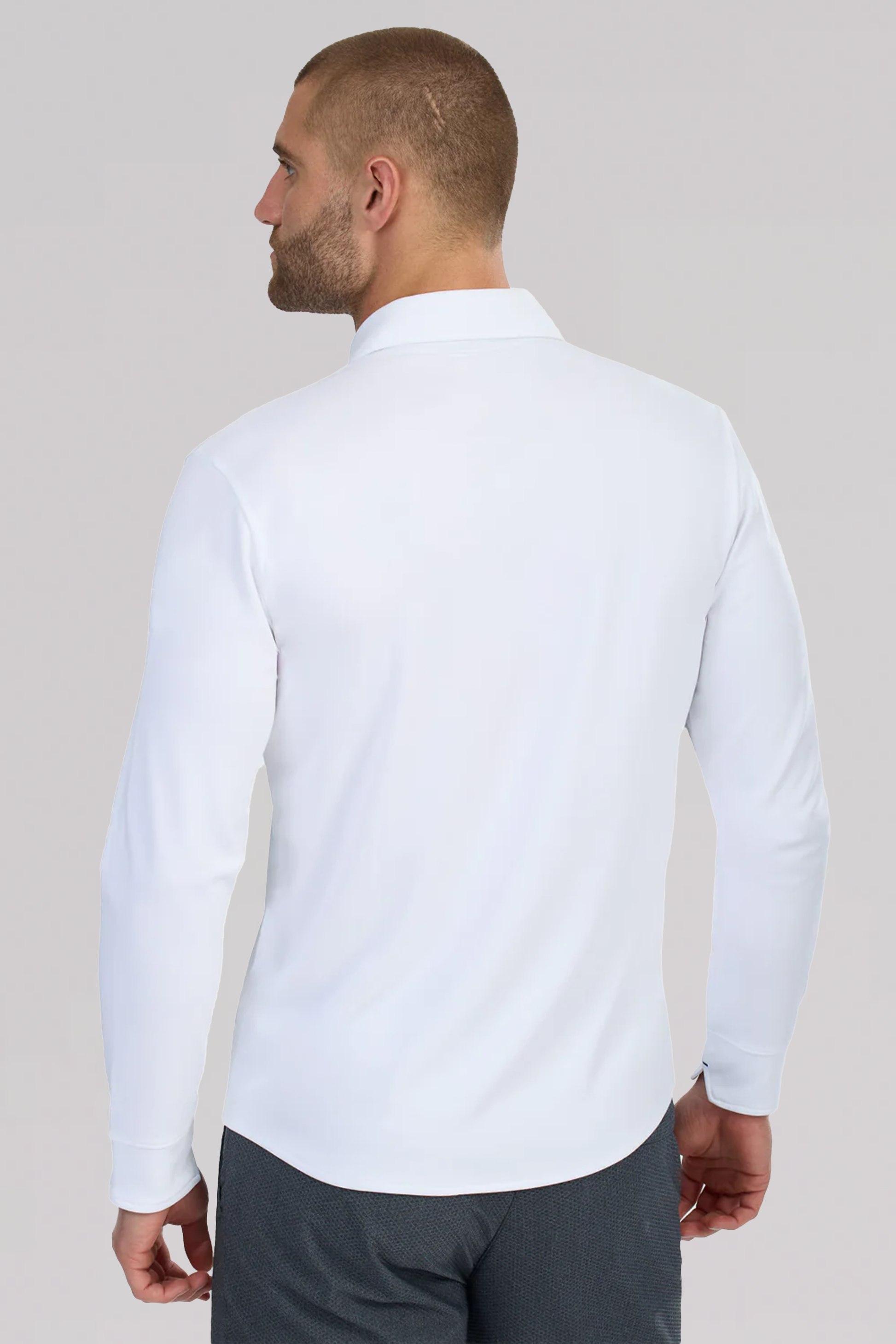 A man with short hair and a beard is seen from the back wearing the Twillory Long Sleeve Button Down Polo Contrast in white with navy stitch. The background is light, and his left arm is slightly bent. He pairs it with dark gray pants.