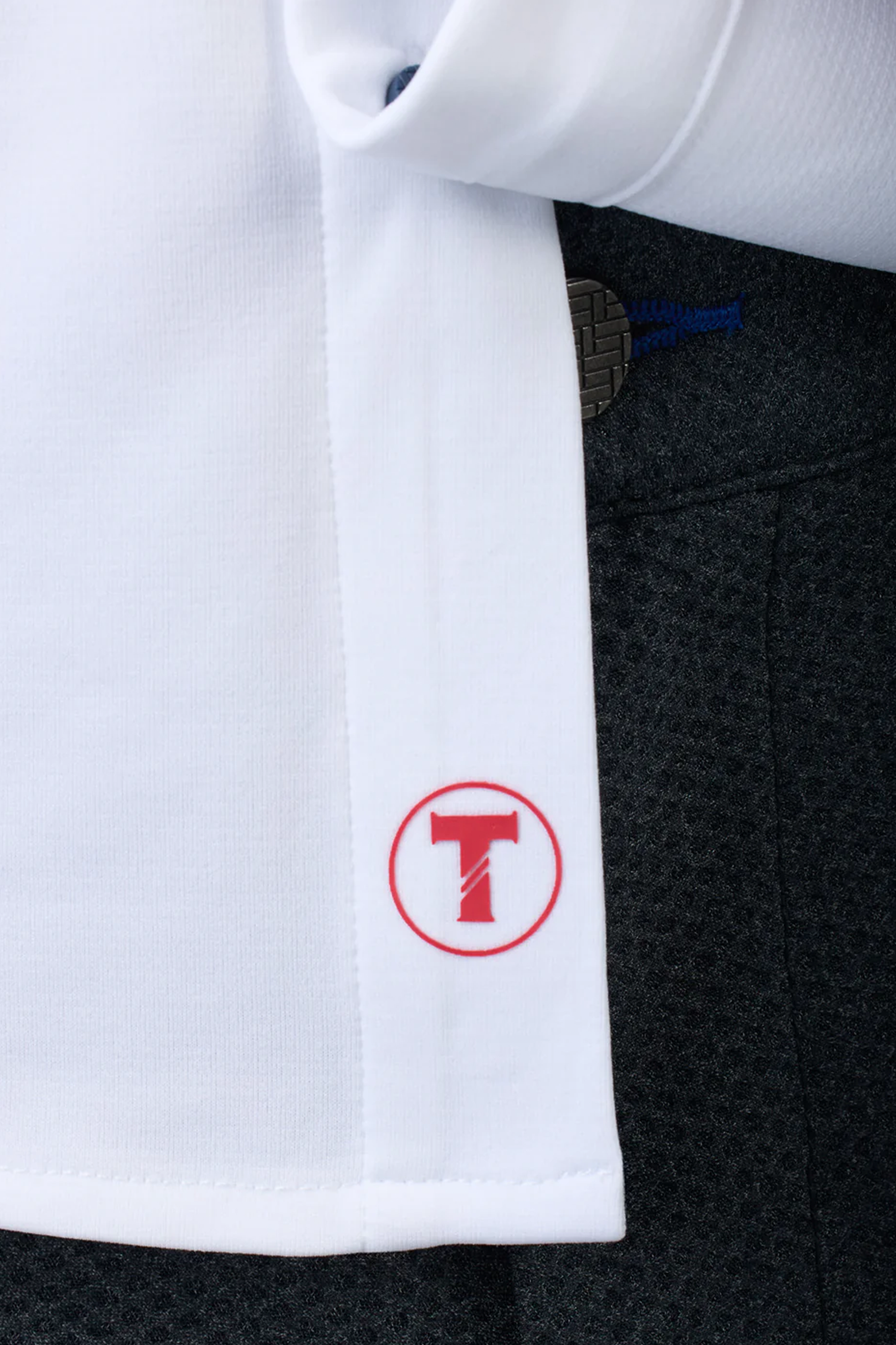 Close-up of Twillory’s Long Sleeve Button Down Polo Contrast in white with navy stitching, featuring a red “T” logo by the hem. The smooth, moisture-wicking fabric is paired with dark-textured pants shown in the background.