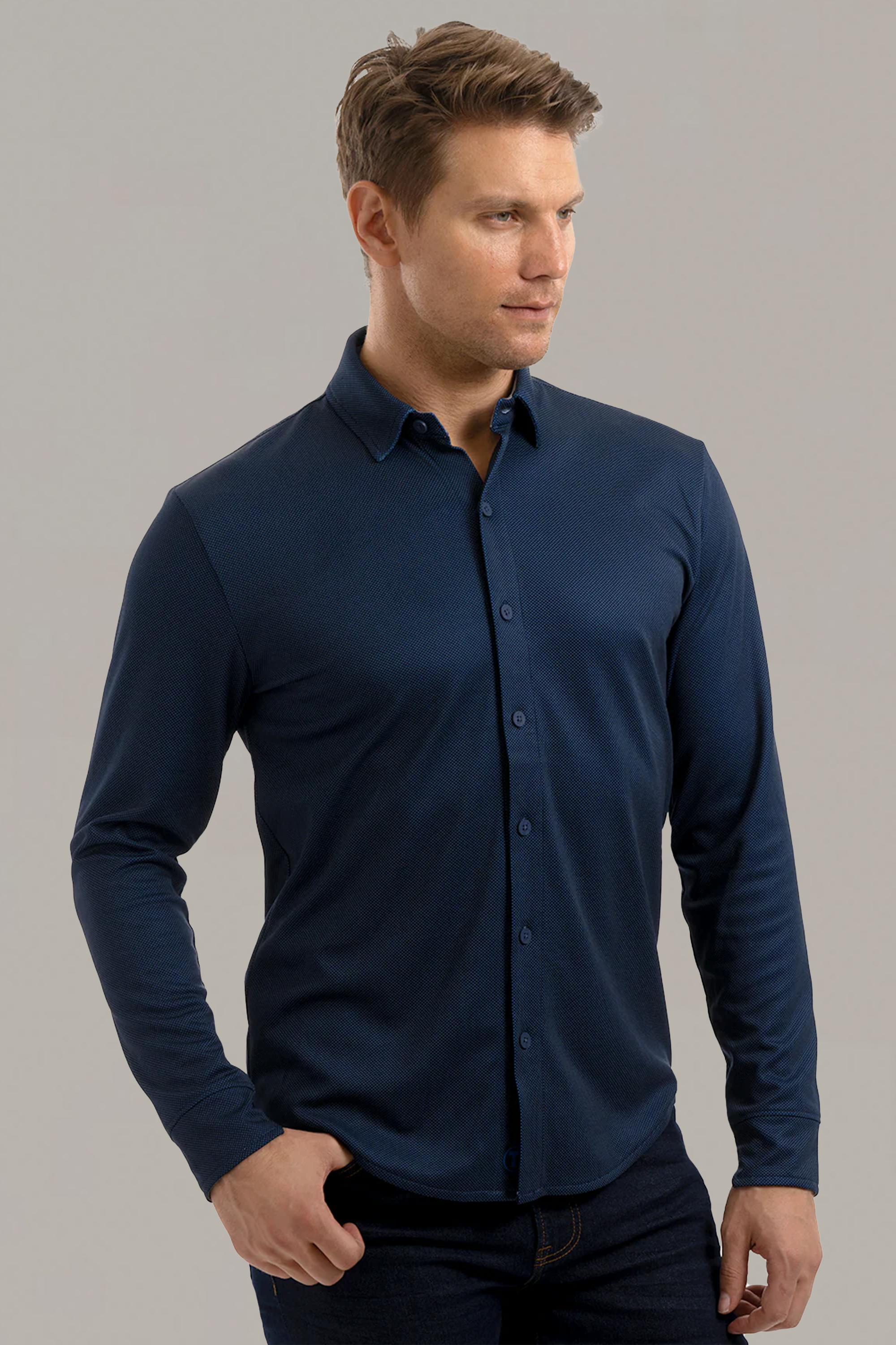A man stands against a light grey background, wearing the Twillory Long Sleeve Button Down Polo Dot in Navy with dark jeans. He looks slightly right with a neutral expression, one hand in his pocket and neatly styled brown hair.