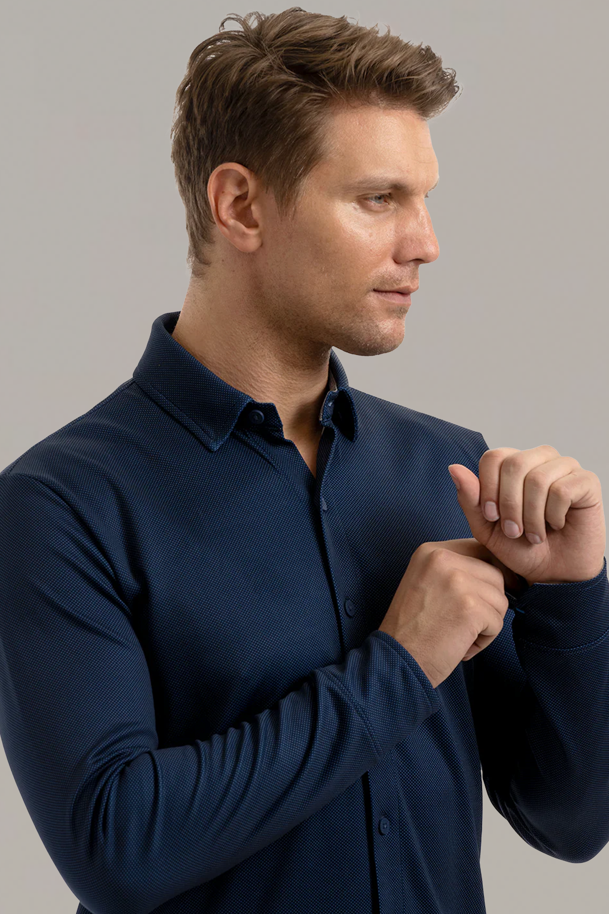 A man with short light brown hair wears the Twillory Long Sleeve Button Down Polo Dot in Navy. He stands in profile against a light background, with hands near his chest as if adjusting the shirt, and displays a neutral expression.