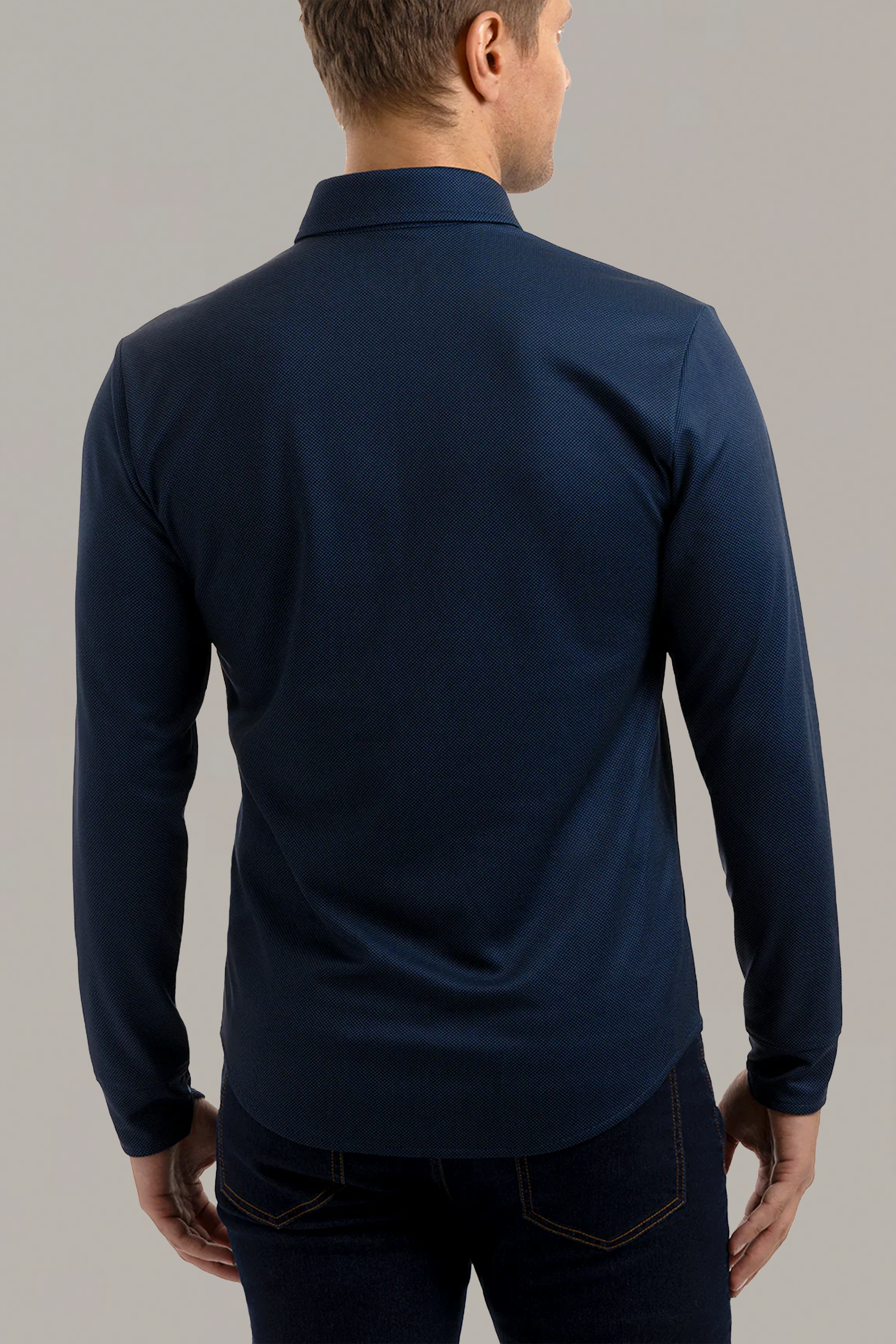 A man faces away from the camera, wearing the Twillory Long Sleeve Button Down Polo Dot in Navy with dark jeans. His arms at his sides display the plain back of the collared shirt, which features no visible logos or patterns.