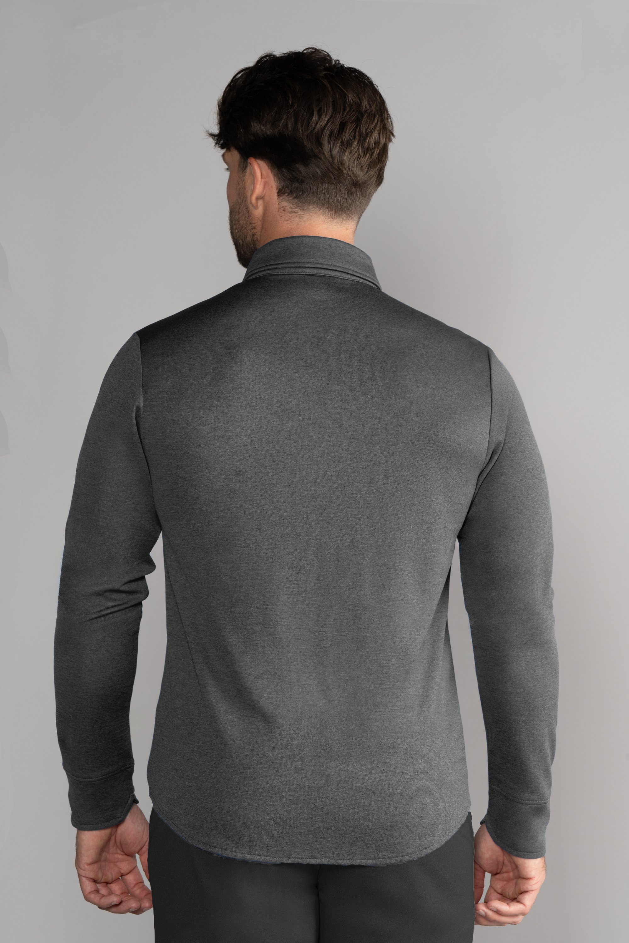 A man with short brown hair is shown from behind, wearing Twillory’s Long Sleeve Button Down Polo Solid in Charcoal, standing against a plain light gray background with his arms relaxed by his sides.