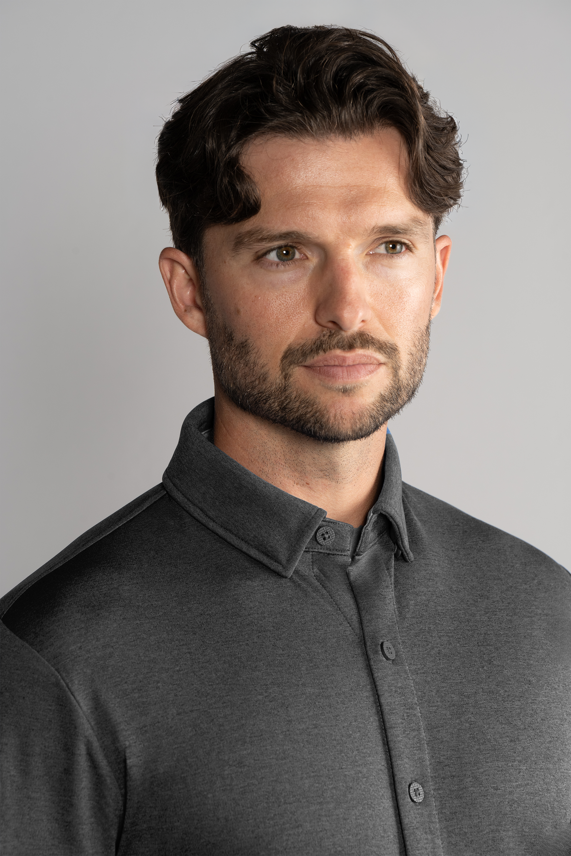 A man with wavy brown hair, a trimmed beard, and light skin stands against a plain light gray background. He wears the Twillory Long Sleeve Button Down Polo Solid in Charcoal and looks slightly to the right with a neutral, thoughtful expression.