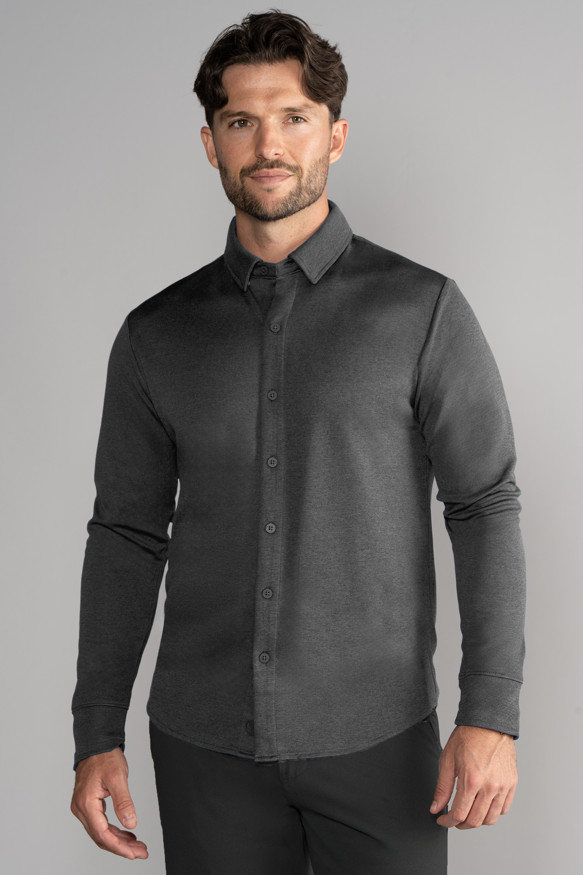 A man with short brown hair and a trimmed beard wears the Twillory Long Sleeve Button Down Polo Solid in Charcoal, paired with black pants. He stands facing forward against a light gray background, his left arm slightly bent and expression neutral.