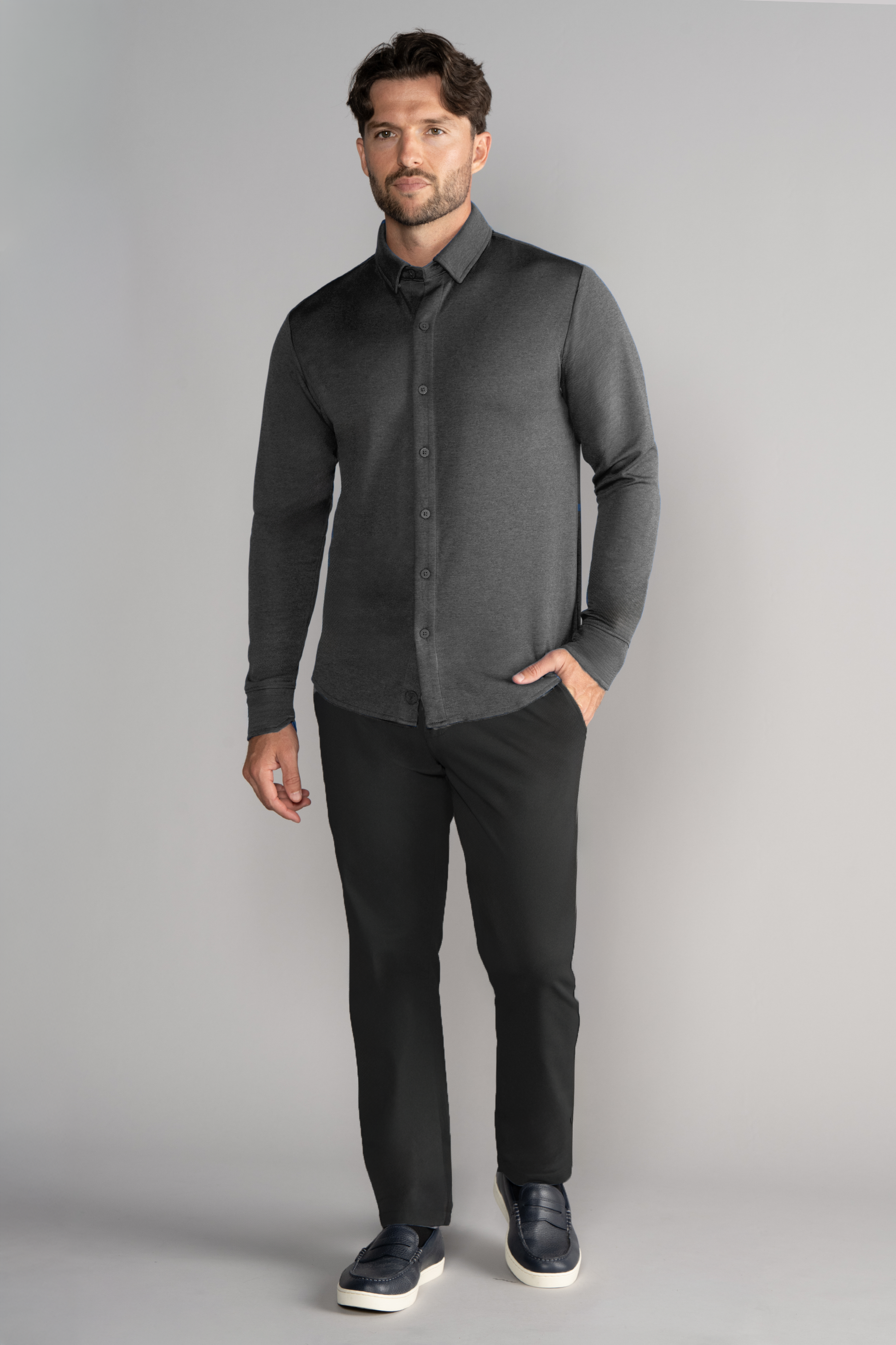 A man stands against a light gray backdrop, wearing the Twillory Long Sleeve Button Down Polo Solid in Charcoal. He has one hand in his pocket, faces forward, and pairs the shirt with black pants and dark slip-on shoes.