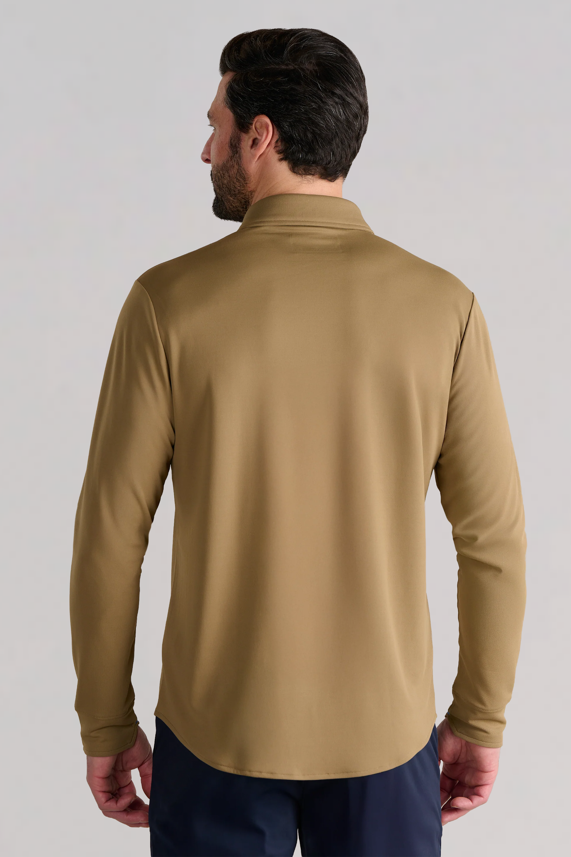 A man with short dark hair and a beard is shown from the back wearing the Twillory Long Sleeve Button Down Polo Solid in Tobacco with dark pants, standing against a light grey background with his arms relaxed at his sides.