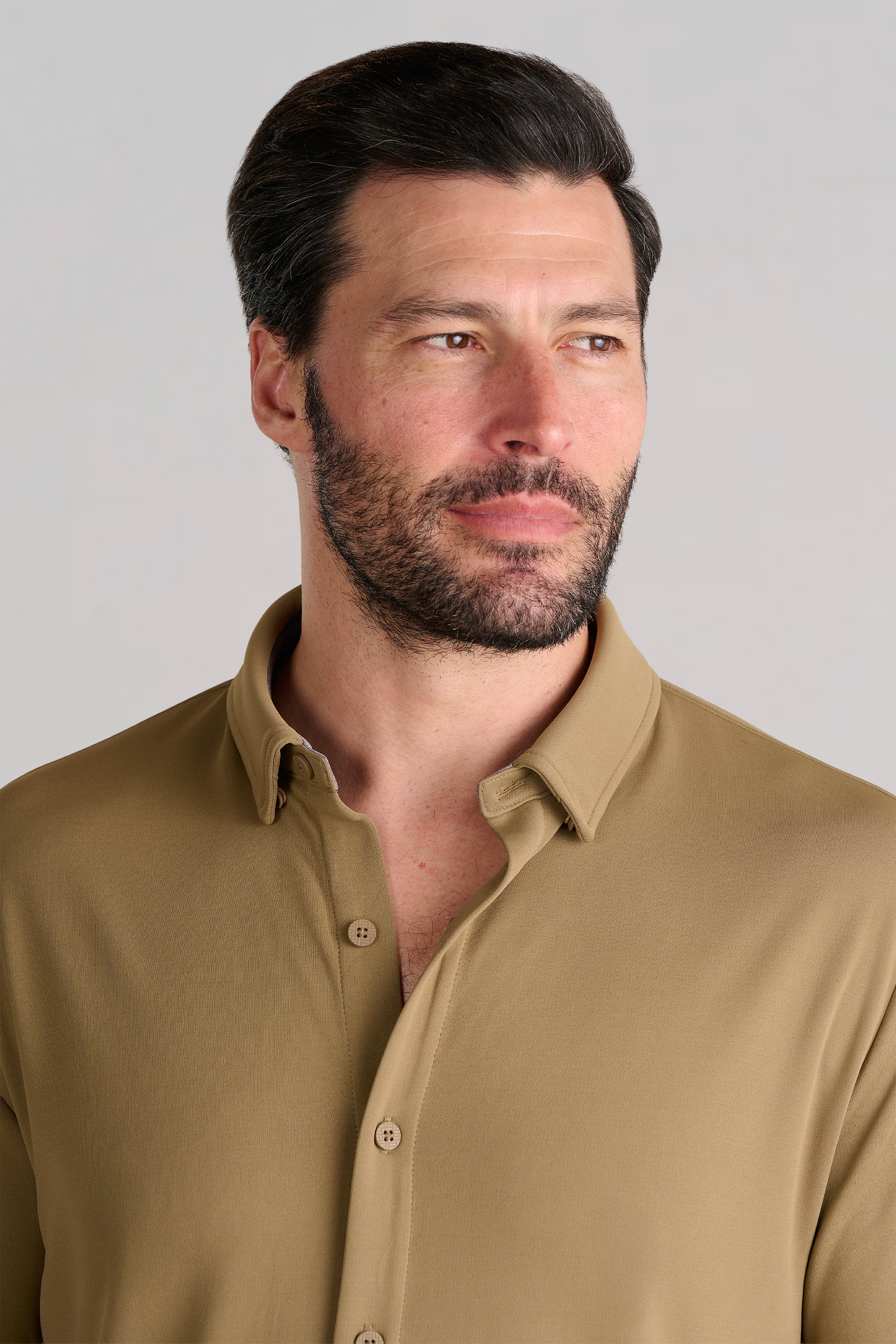 A man with short dark hair and a full beard wears the Twillory Long Sleeve Button Down Polo Solid in Tobacco. He looks slightly right, neutral-faced, against a plain light gray background with only his upper torso visible.