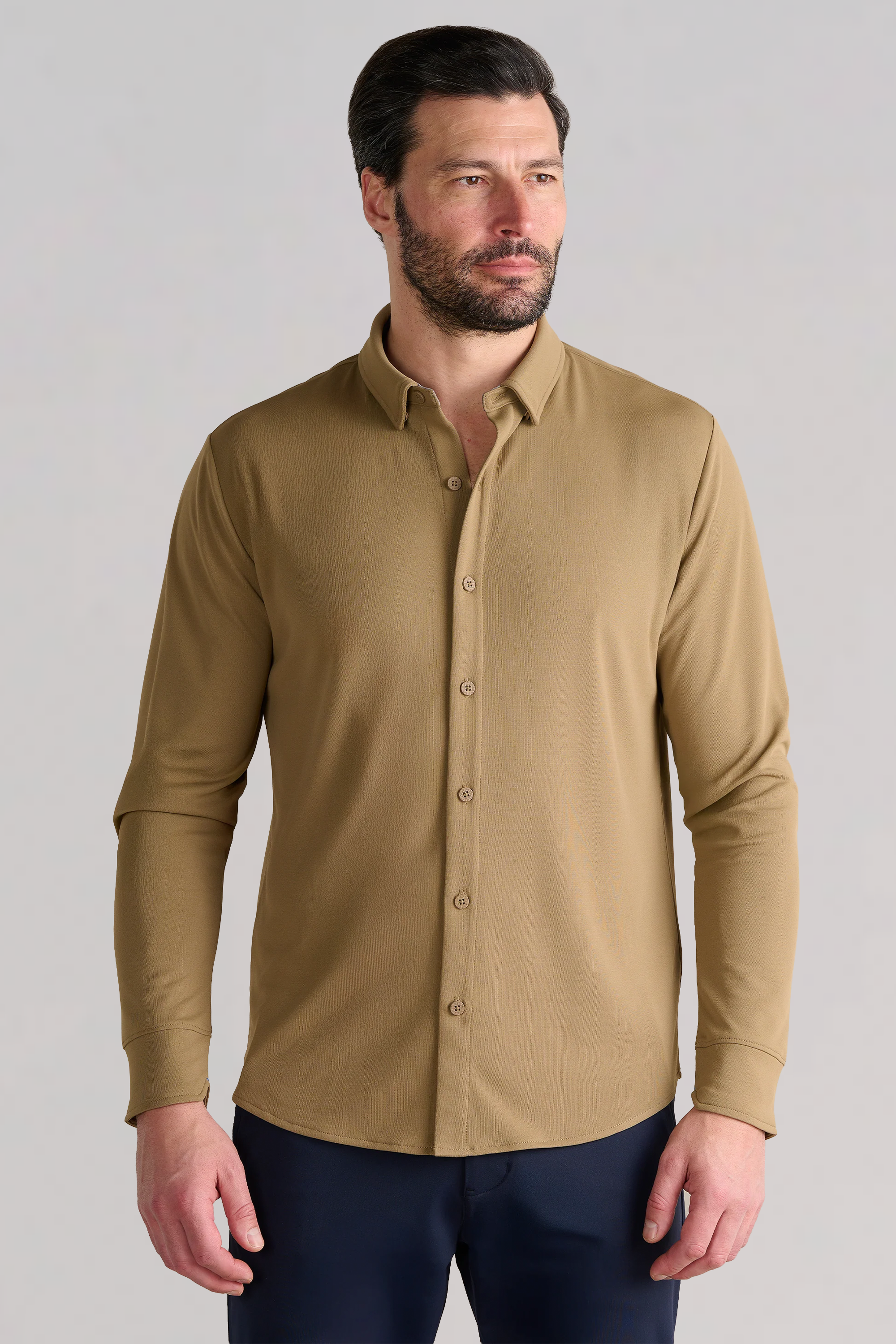 A man with short dark hair and a beard wears the Twillory Long Sleeve Button Down Polo Solid in Tobacco, standing slightly turned right with arms relaxed against a plain light gray background.