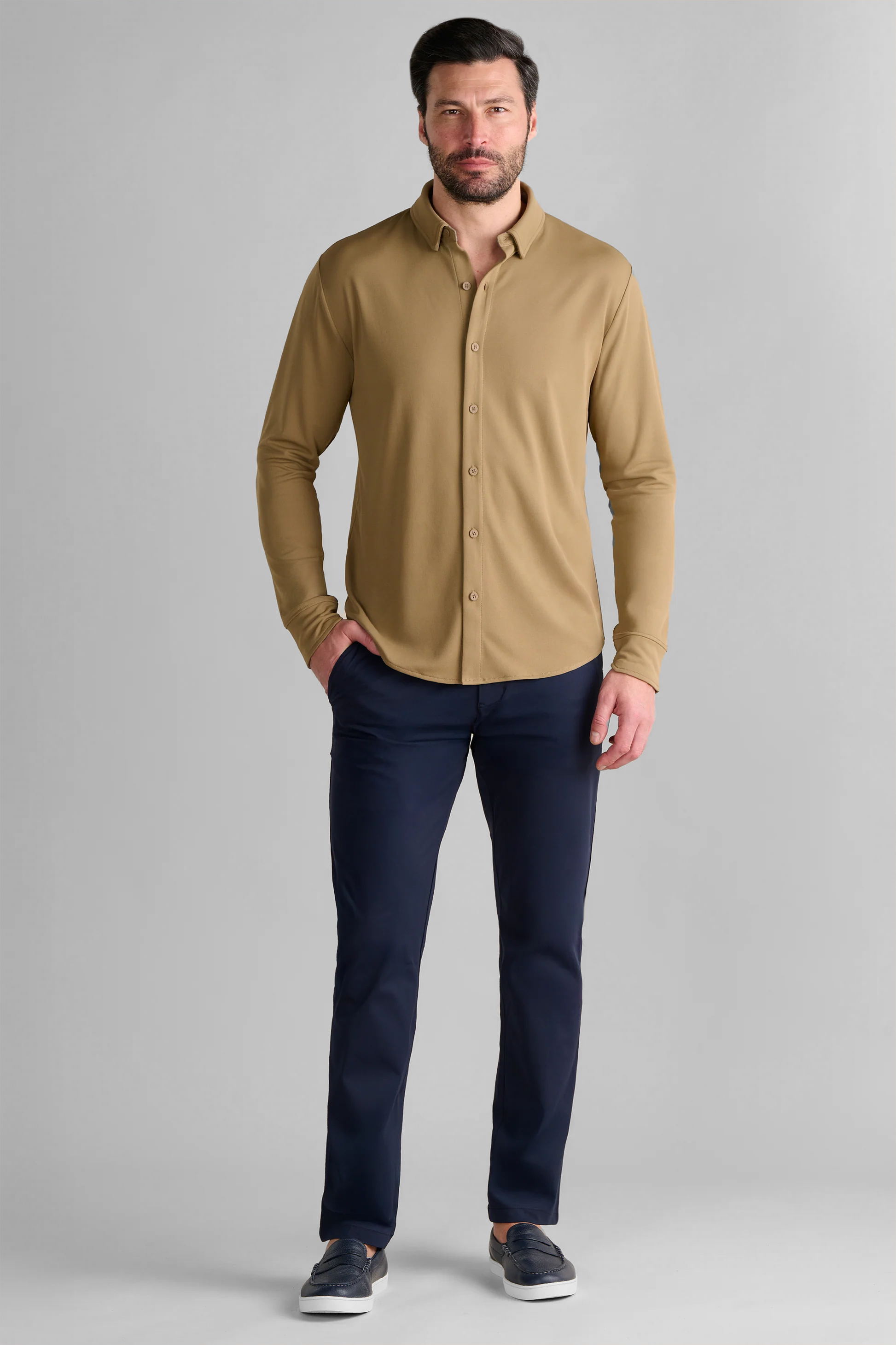 A man with short dark hair and a trimmed beard stands against a light gray background, wearing the Twillory Long Sleeve Button Down Polo Solid in Tobacco, paired with navy pants and dark slip-on shoes. One hand rests in his pocket; his expression is neutral.