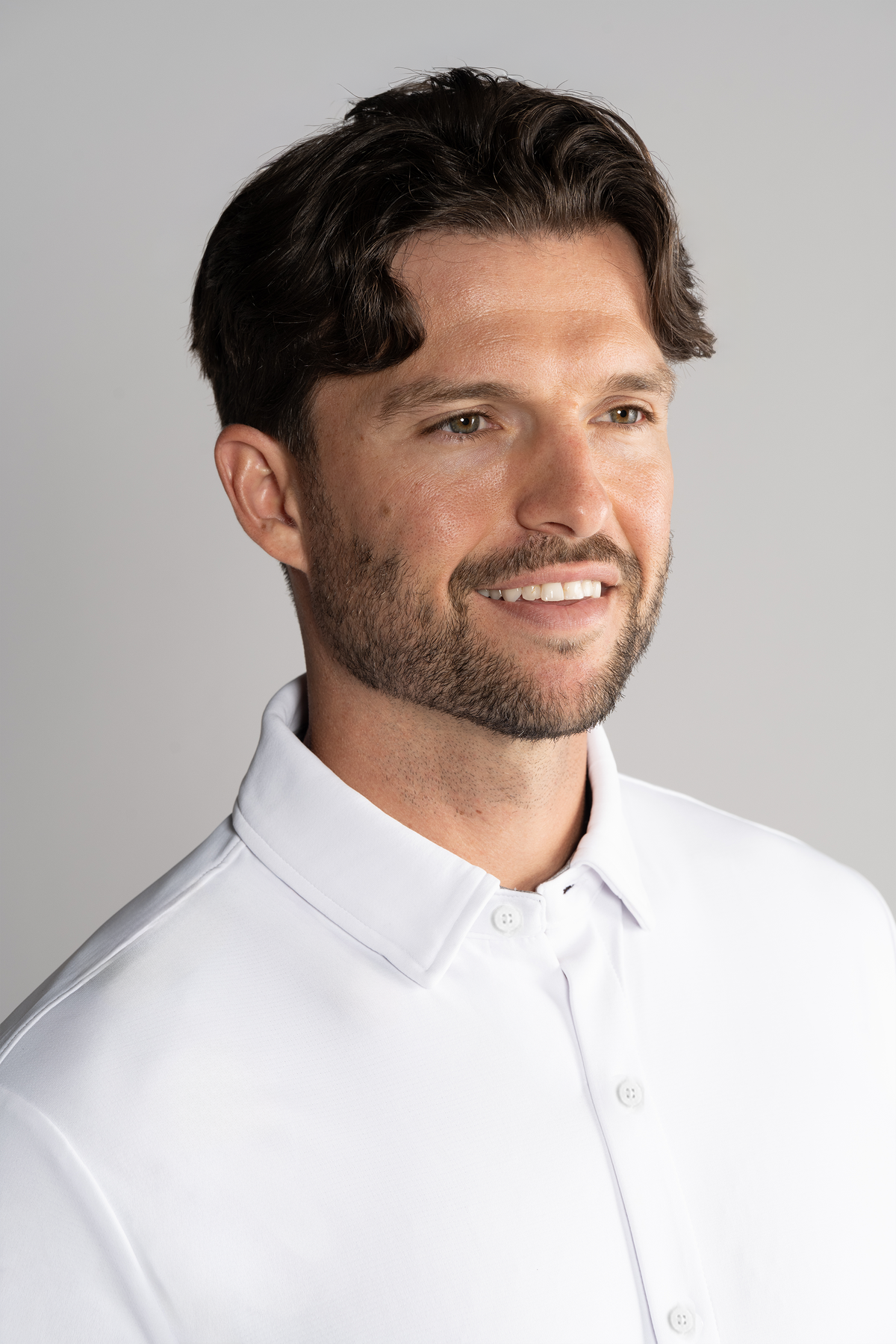 A man with short, dark, wavy hair and a trimmed beard smiles while looking to the side, wearing Twillory’s Long Sleeve Button Down Polo Solid in White against a plain light gray background with soft lighting.