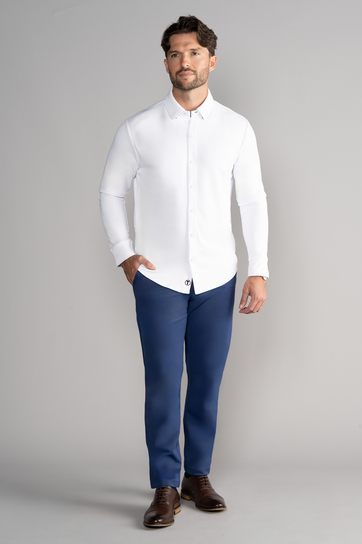 A man with short brown hair and a beard stands against a light gray background, wearing the Twillory Long Sleeve Button Down Polo Solid in White. He pairs it with blue pants and brown dress shoes, facing forward with a neutral expression.