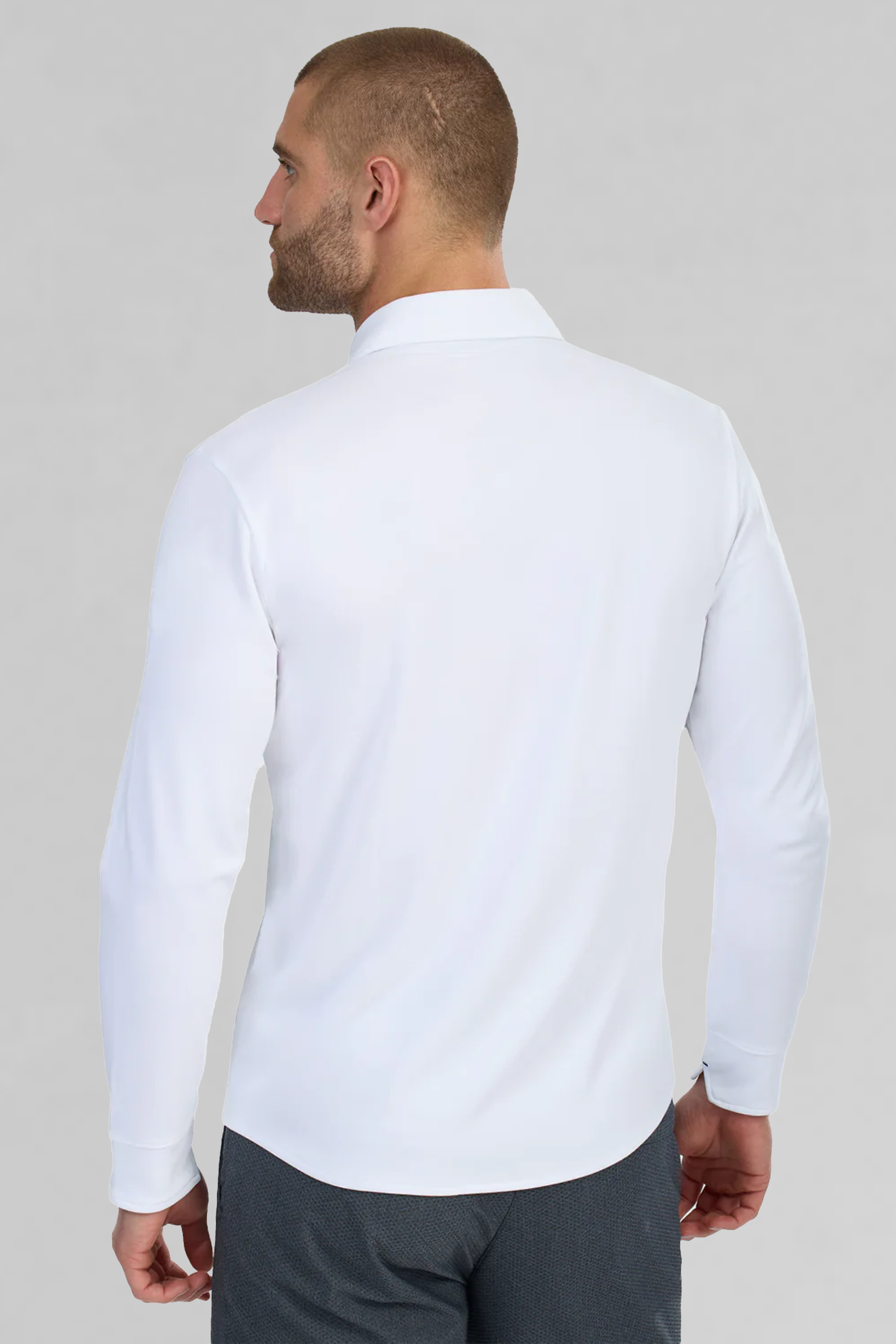 A man with short hair and a beard stands with his back to the camera, wearing Twillory’s Long Sleeve Button Down Polo Contrast in Powder Blue White. The plain, light gray background highlights the classic style of this shirt.