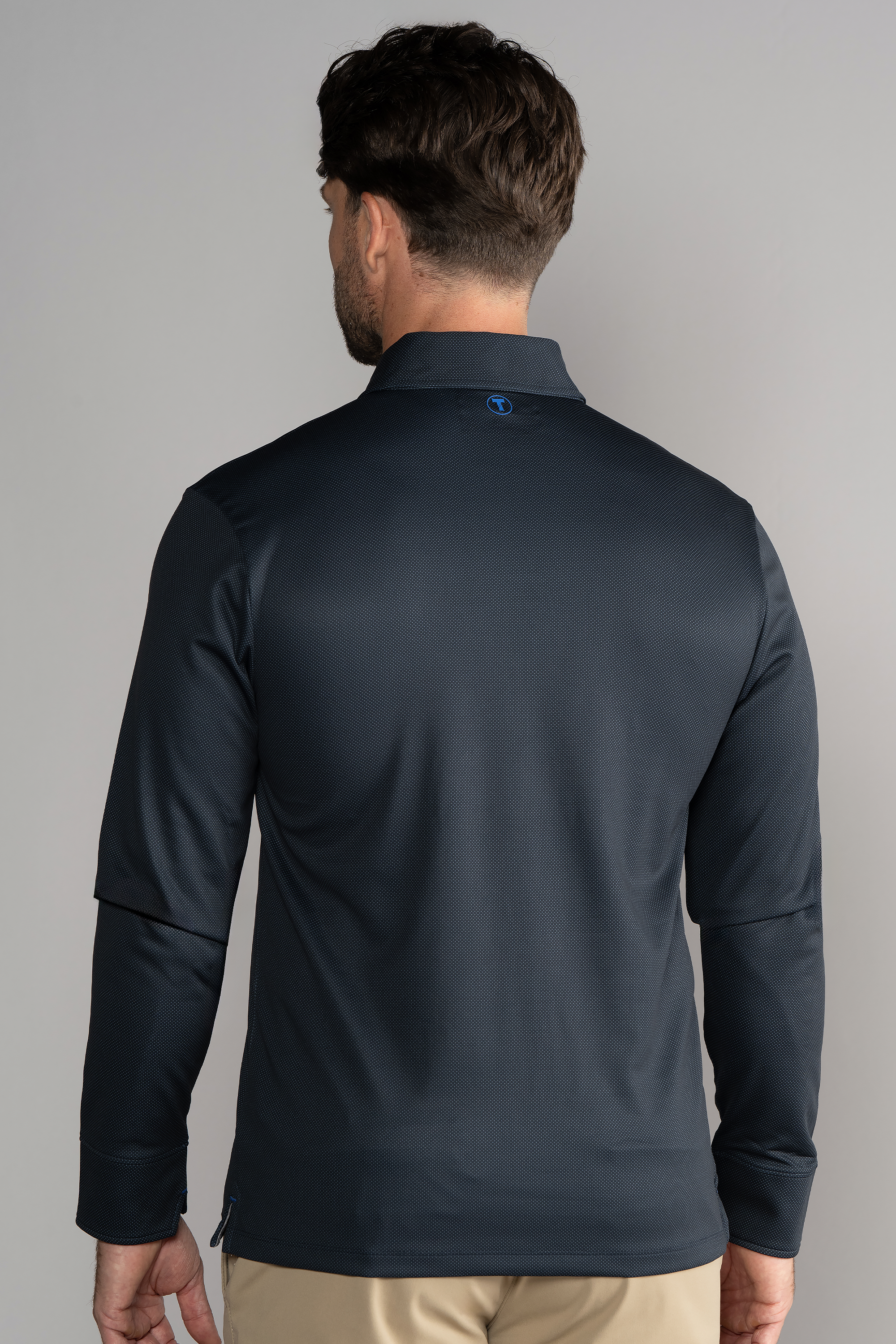 A man with short, dark brown hair is seen from behind wearing a Twillory Long Sleeve Polo Dot in Charcoal and light tan pants, against a plain light gray background. The shirt features a small blue logo at the back of the collar.