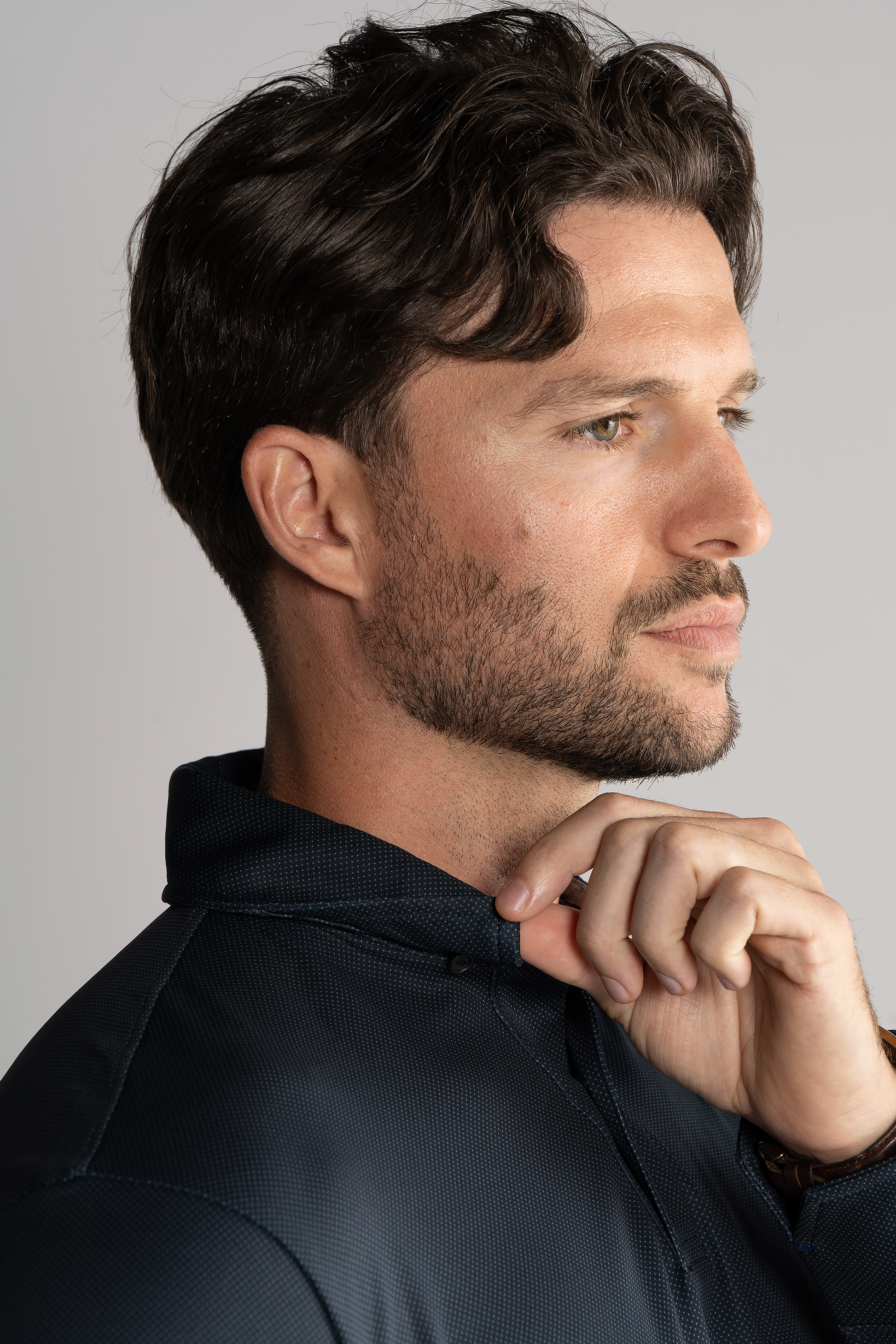 A man with short, wavy dark hair and a trimmed beard faces right in profile, wearing the Twillory Long Sleeve Polo Dot in Charcoal. He gently holds the collar with his right hand, his thoughtful expression set against a plain background.