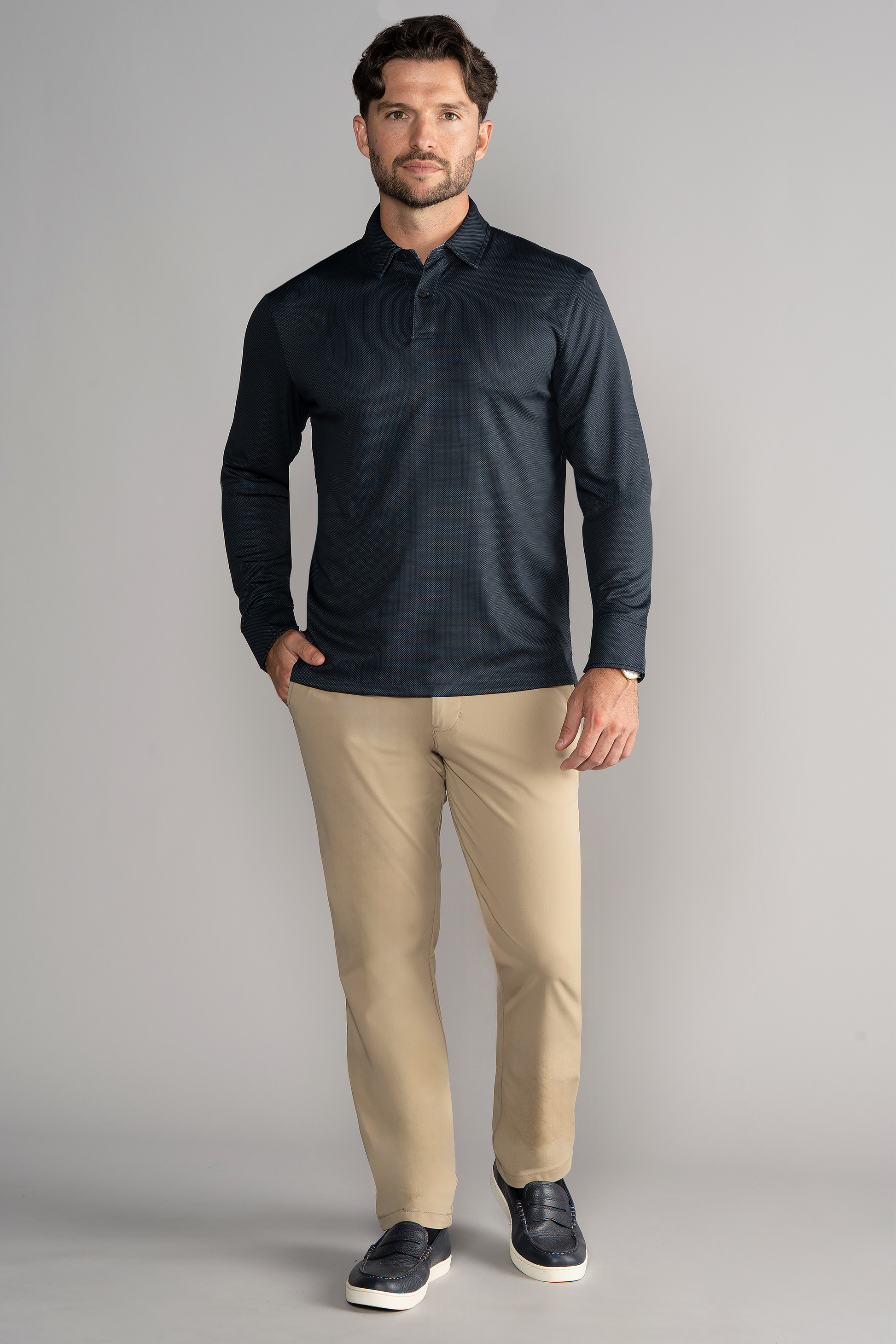 A man with short brown hair and a beard stands against a gray background wearing Twillory's Long Sleeve Polo Dot in Charcoal, beige pants, and black slip-on shoes. His left hand is partially in his pocket as he looks ahead, slightly smiling.