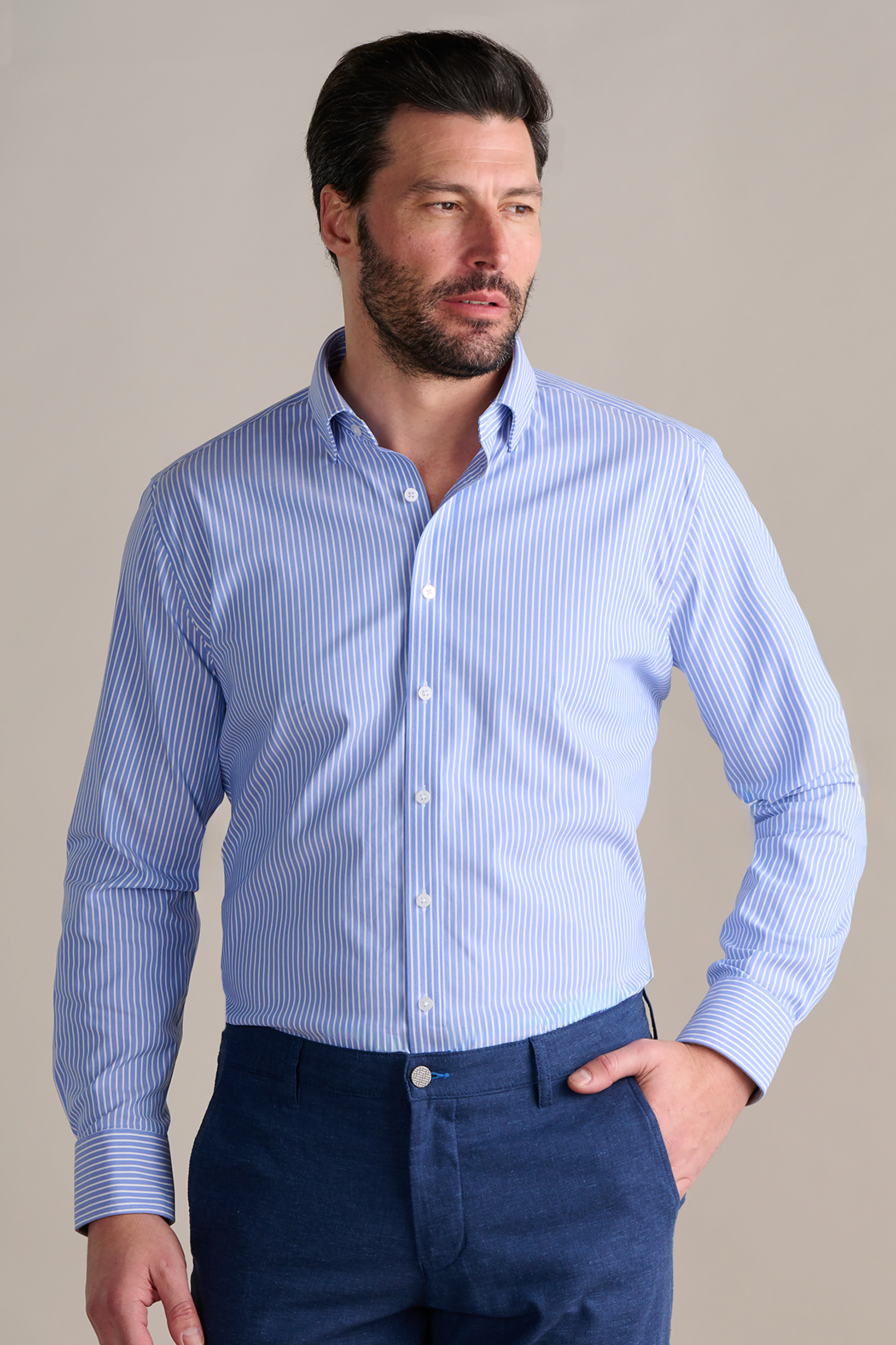 A man with dark hair and a beard wears the Twillory Leader Stripe // Mini Blue Performance Shirt, featuring light blue and white vertical stripes. The shirt is tucked into dark blue trousers as he stands against a beige background, looking slightly right.