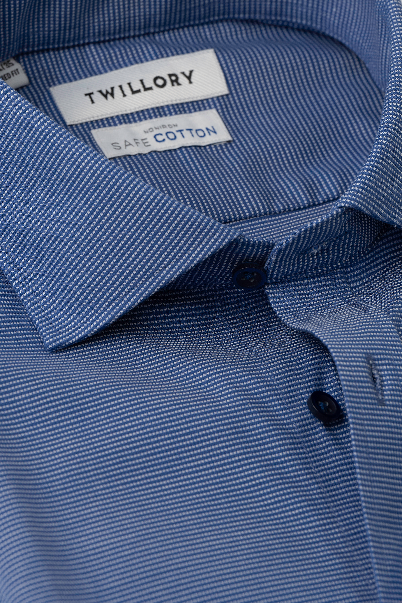 Close-up of Twillory's Non-Iron Twilight Pin Dot shirt in blue, featuring a fine white microstripe. The partially buttoned, hyperbreathable shirt reveals dark blue buttons and inside-collar labels reading Twillory and SafeCotton.