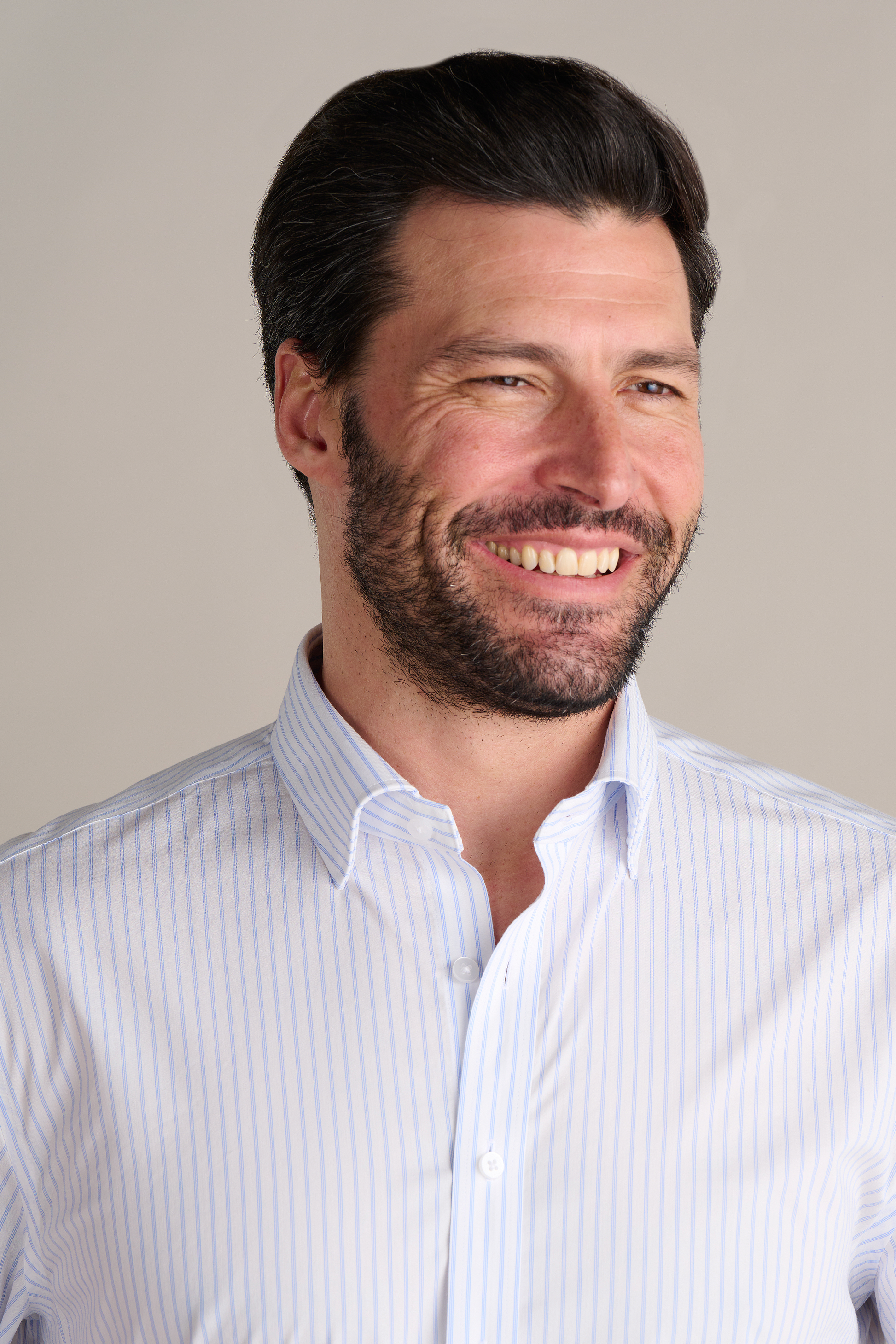 A man with dark hair and a beard smiles while looking to the side, wearing Twillory's Leader Stripe // Thick White shirt. The light background accentuates his professional, friendly look and the shirt’s 4-way stretch performance fabric.