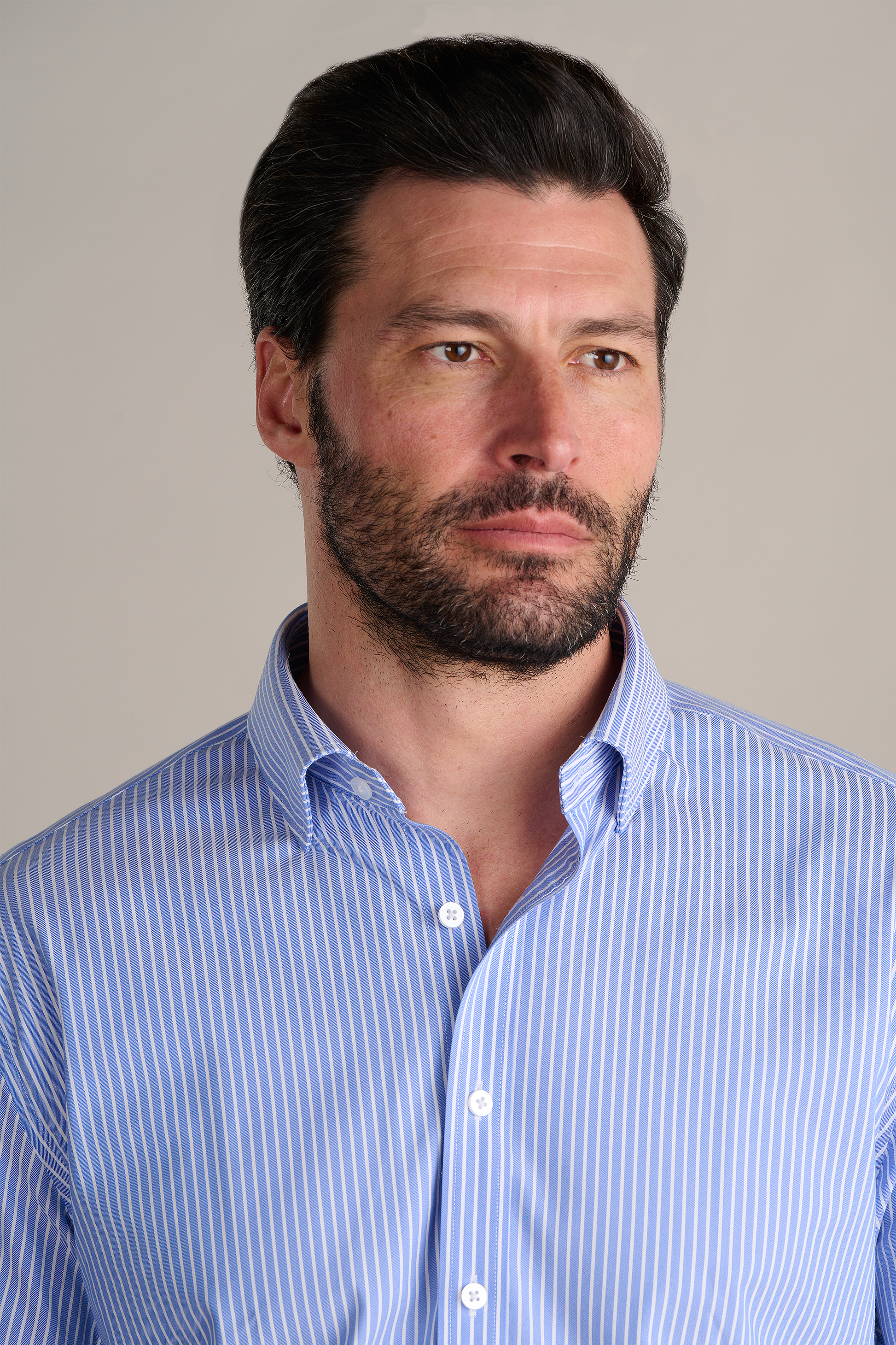 A man with short dark hair and a beard wears Twillory's Leader Stripe // Mini Blue Performance Shirt, featuring light blue and white vertical stripes, against a beige background under soft lighting. He looks slightly left with a neutral expression.