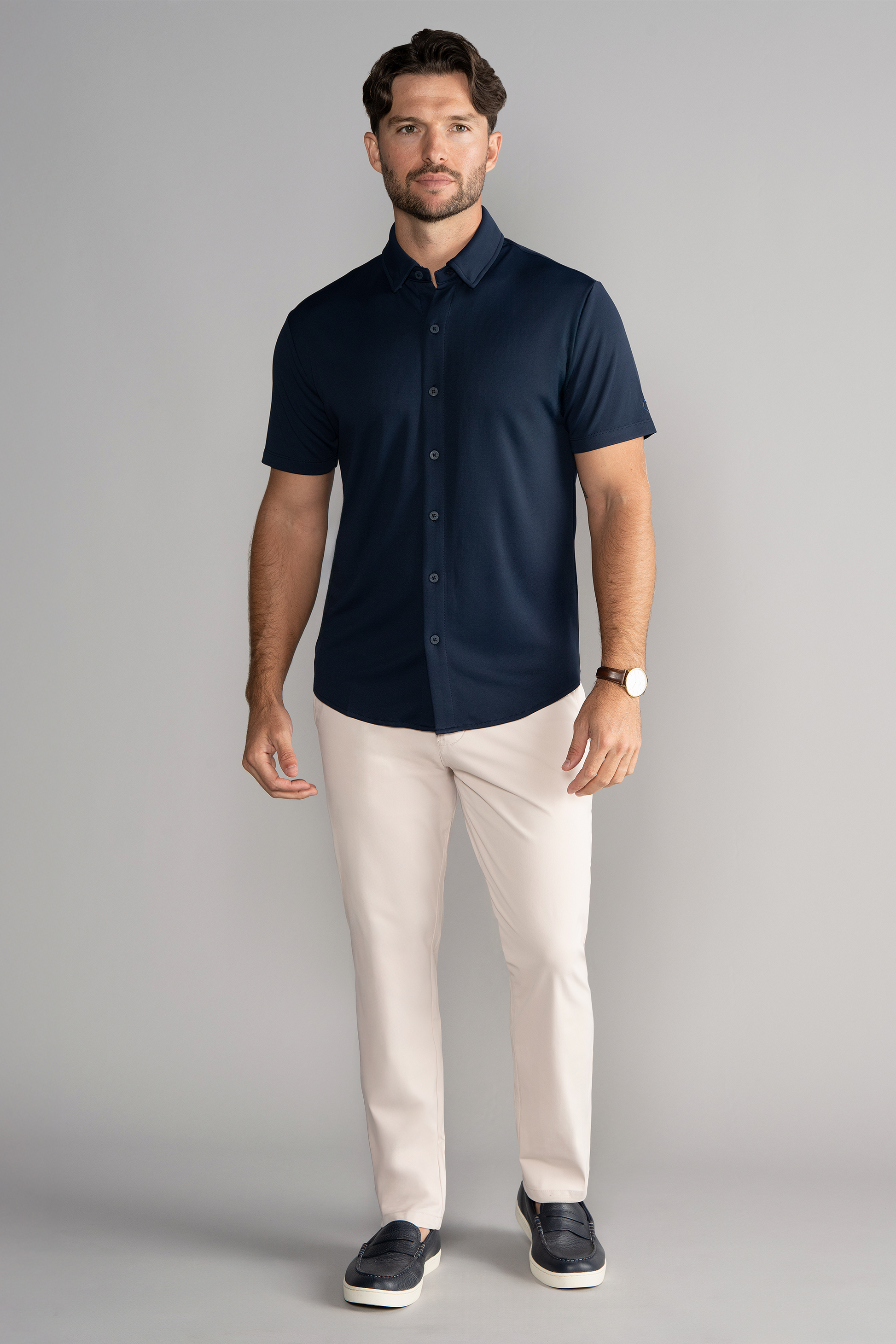 A man with short dark hair and a beard poses neutrally against a gray backdrop, wearing the Twillory Short Sleeve Button Down Polo Solid in Navy, light beige pants, black slip-on shoes, and a wristwatch on his left wrist.