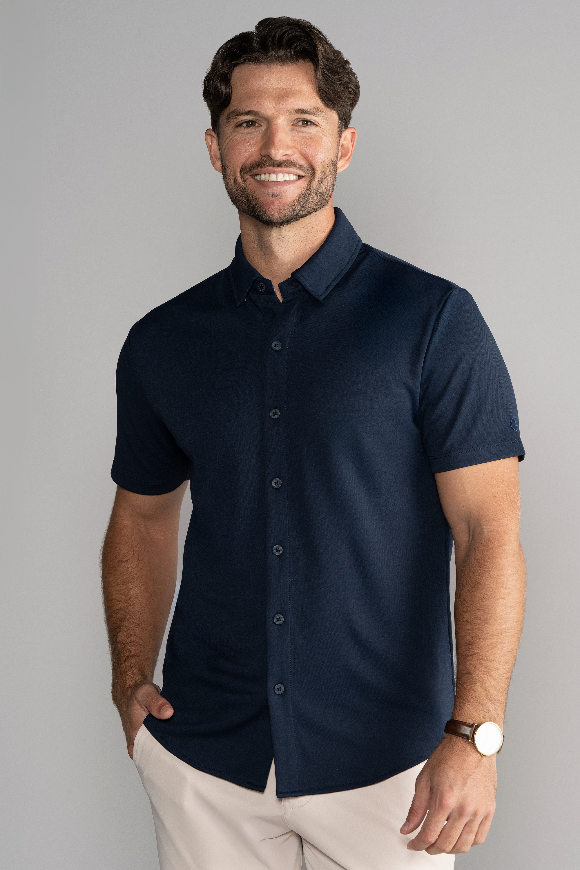 A man with wavy brown hair and a short beard smiles against a light gray background. He wears the Twillory Short Sleeve Button Down Polo Solid in Navy, beige pants, and a brown wristwatch, with his left hand in his pocket.