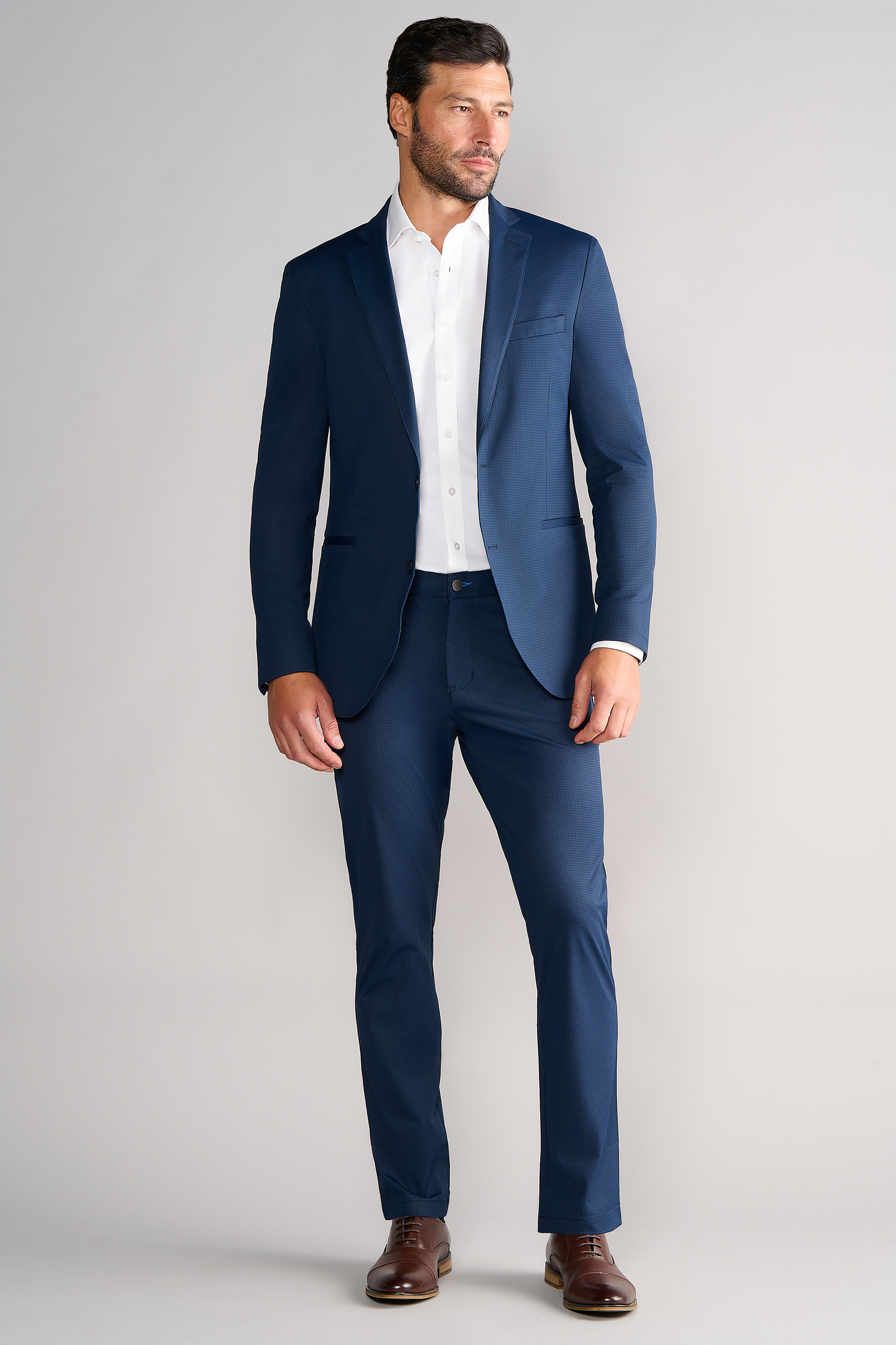 A man with short dark hair and a beard wears the Twillory Performance Blazer in Navy Houndstooth over a white dress shirt and brown dress shoes, standing against a light gray background and looking slightly left with a neutral expression.