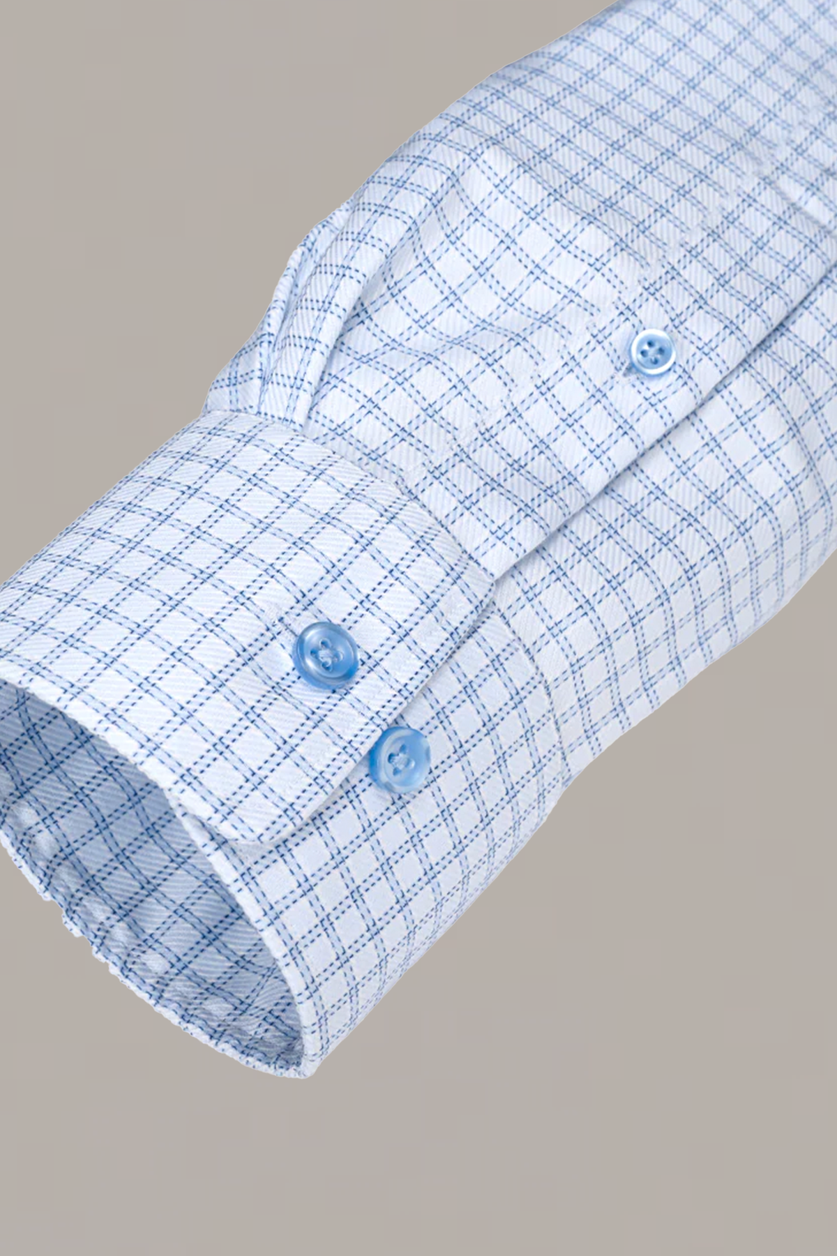 A close-up of Twillory’s Non-Iron Blue Windowpane Twill shirt cuff shows a light blue and white check pattern with two blue buttons on the cuff and one on the placket, made from stretch cotton, set against a light gray background.