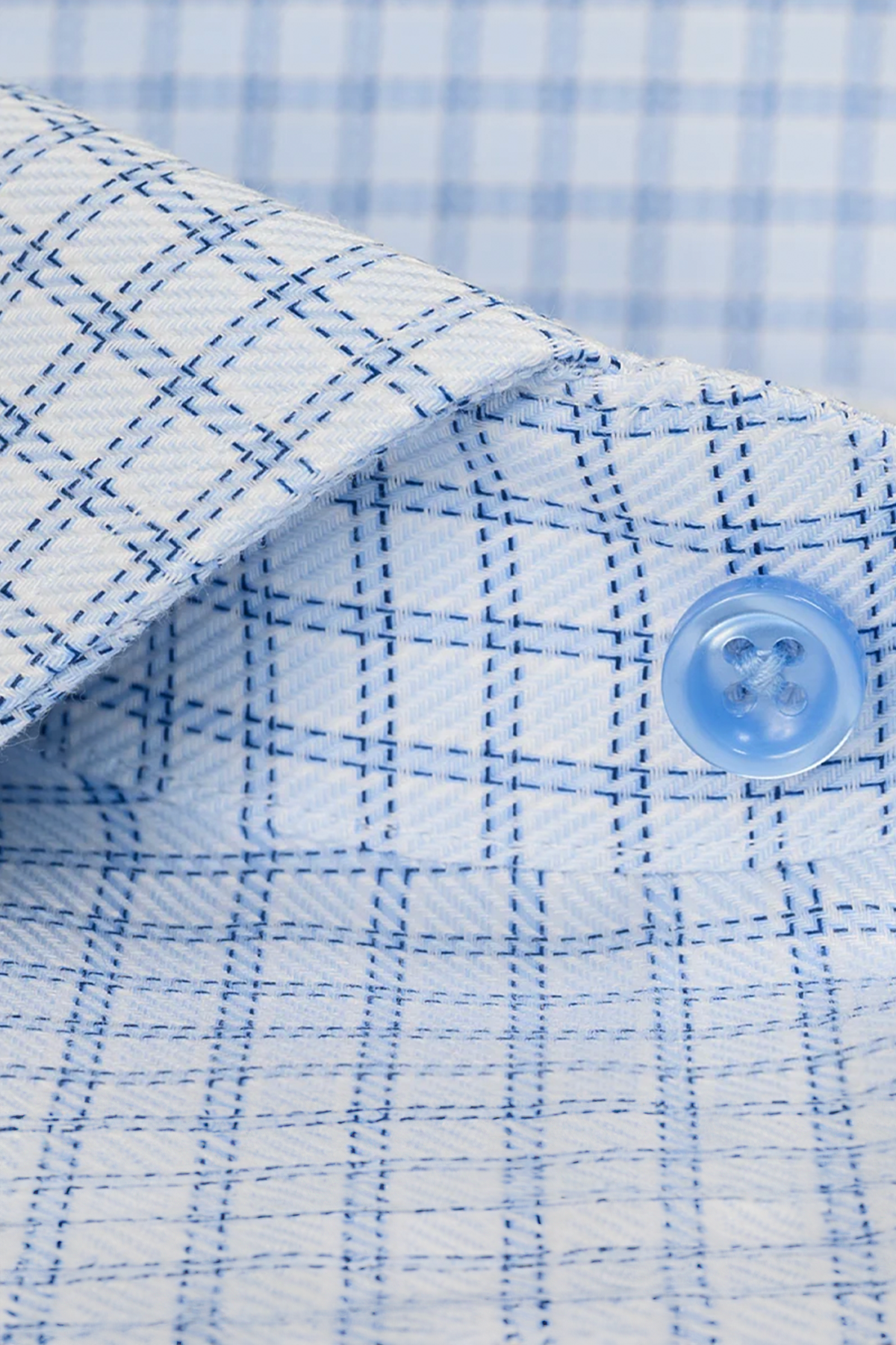 Close-up of Twillory’s Non-Iron Blue Windowpane Twill shirt, featuring a textured light blue and white grid pattern, a pointed collar, and a blue button. The crisp, non-iron fabric is neatly pressed and folded.