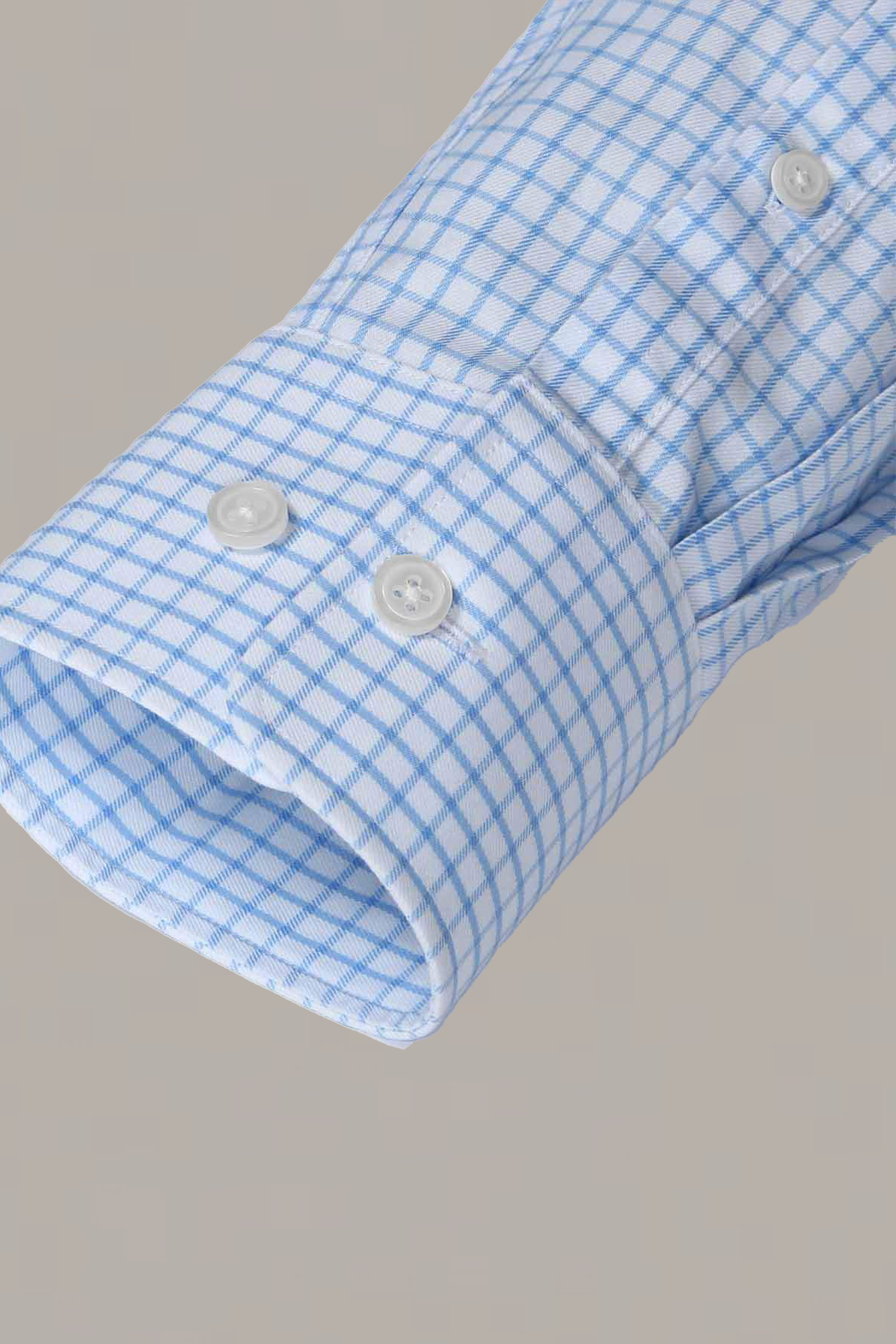 A close-up of the Twillory Non-Iron Sky Windowpane shirt sleeve shows its light blue and white checkered stretch cotton, featuring a double-button cuff with two round white buttons and fine stitching, set against a plain light gray background.